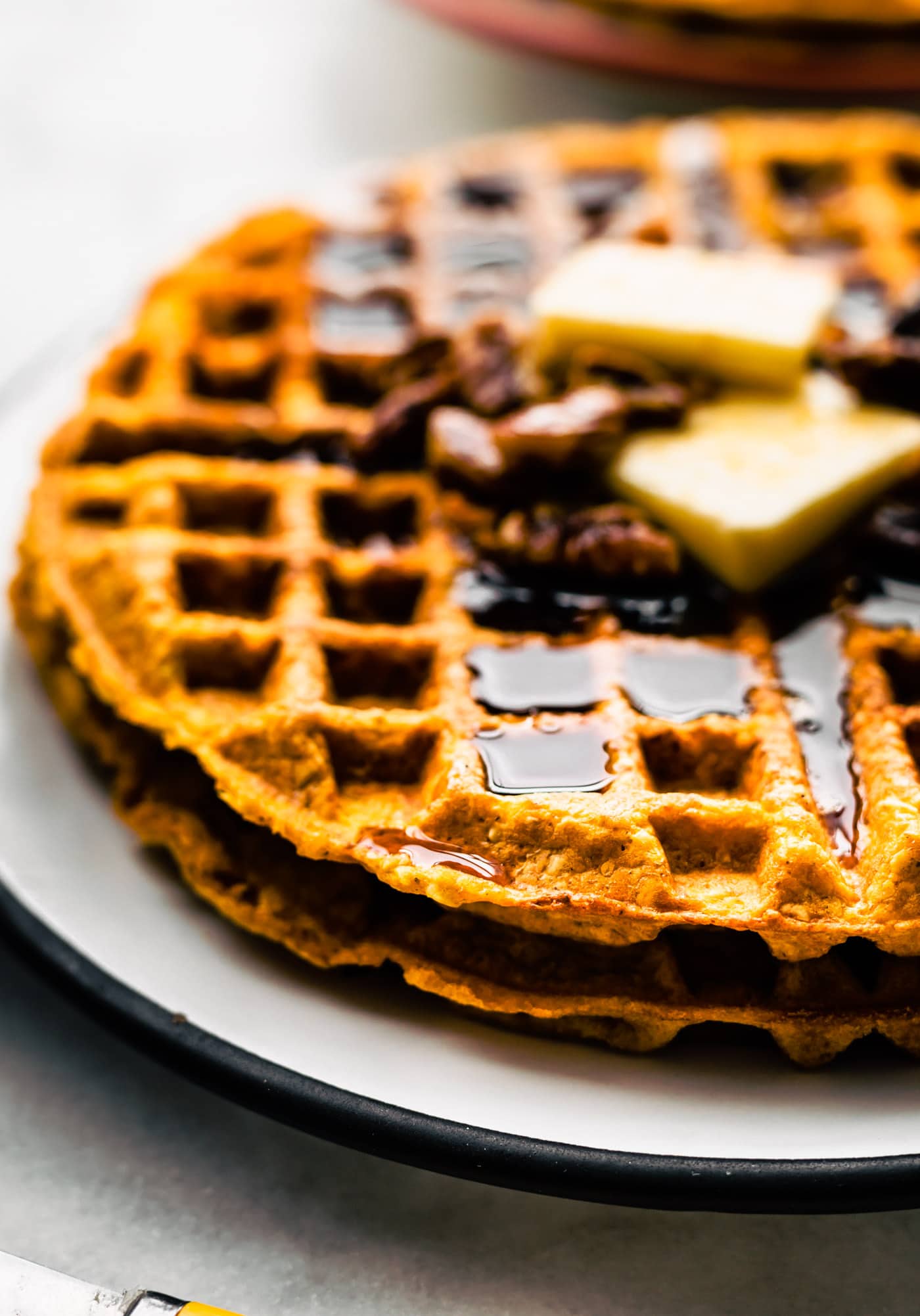 Two round gluten free sweet potato waffles topped with butter and syrup plated on a white plate with a black rim.