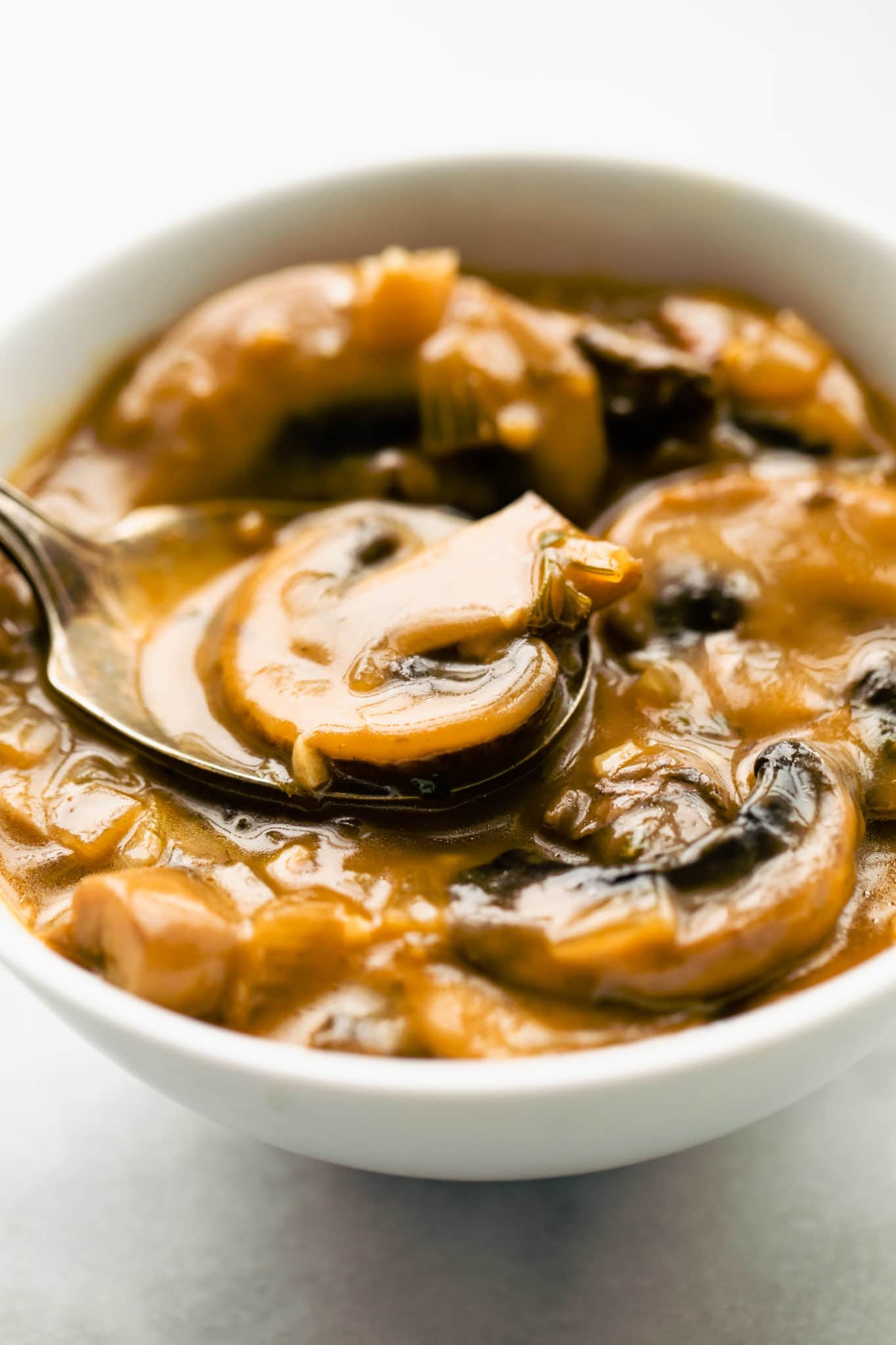 Close up view of a spoon holding a sliced mushroom in gravy over a small white bowl filled with vegan mushroom gravy.