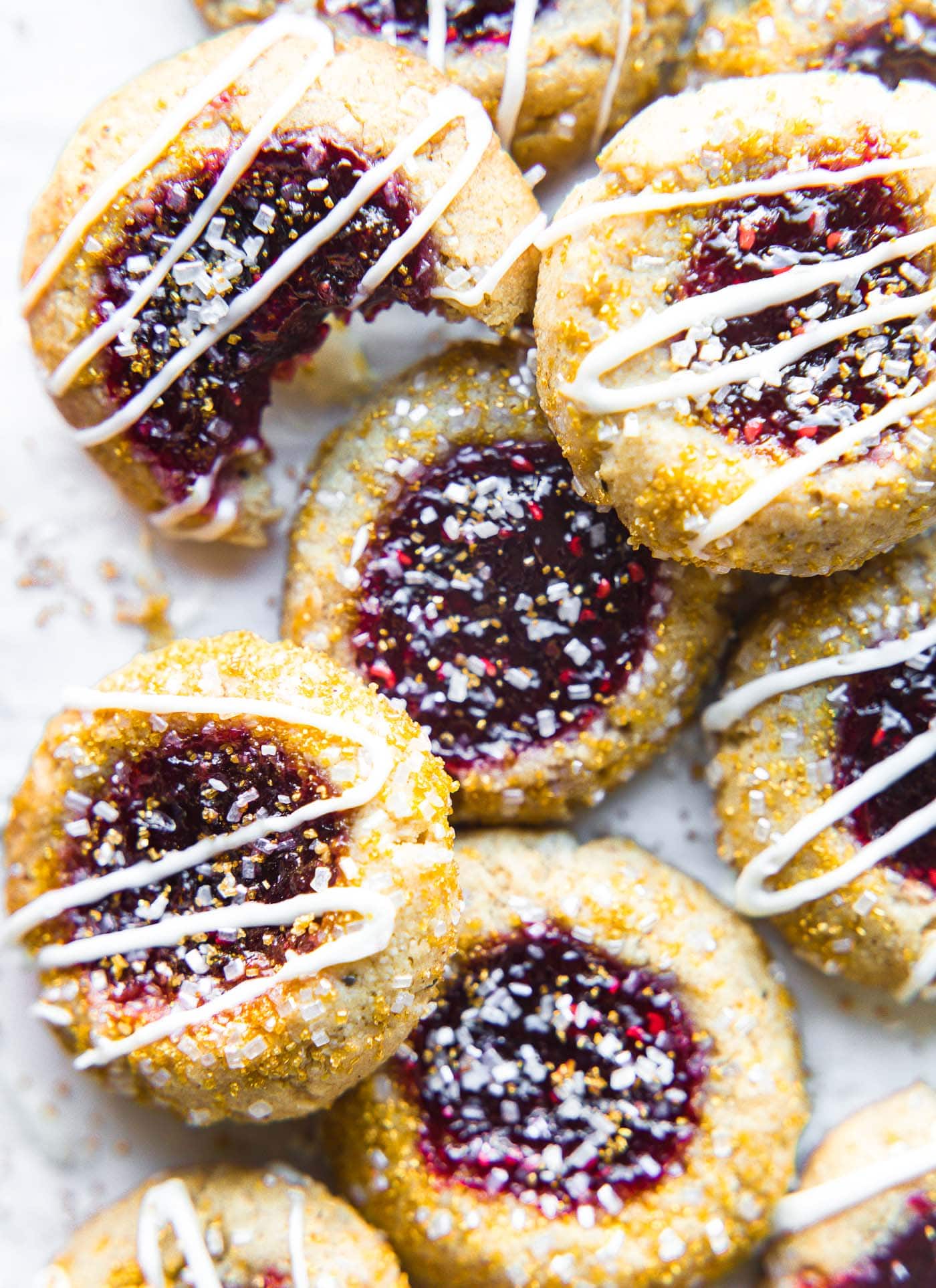 A pile of vegan raspberry thumbprint cookies with a white chocolate drizzle over top and gold sugar crystals sprinkled on top.
