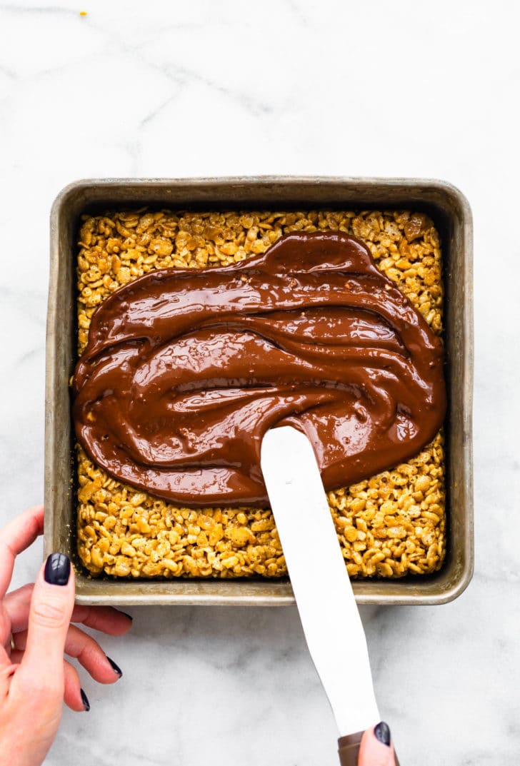 chocolate being spread on top of vegan peanut butter rice krispie treats in a pan