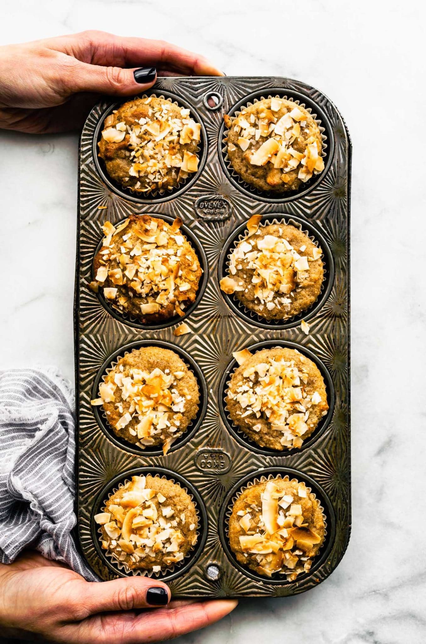 two hands holding a muffin tin full of baked almond flour banana muffins with coconut