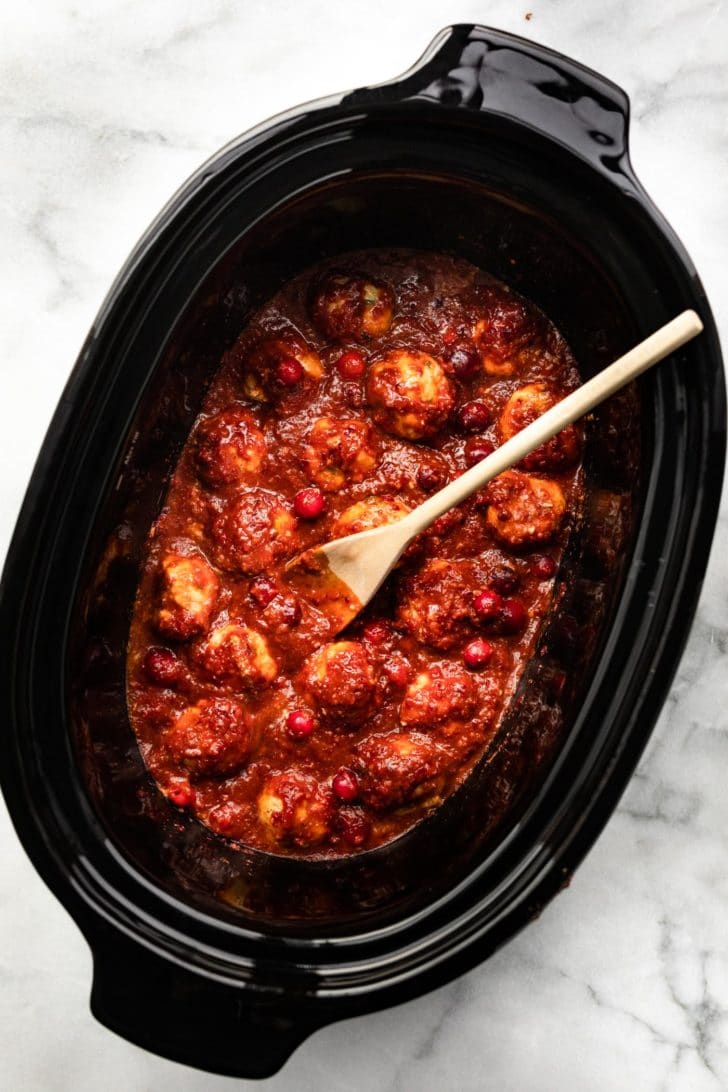 a slow cooker filled with gluten free meatballs with a cranberry sauce with a wooden spoon