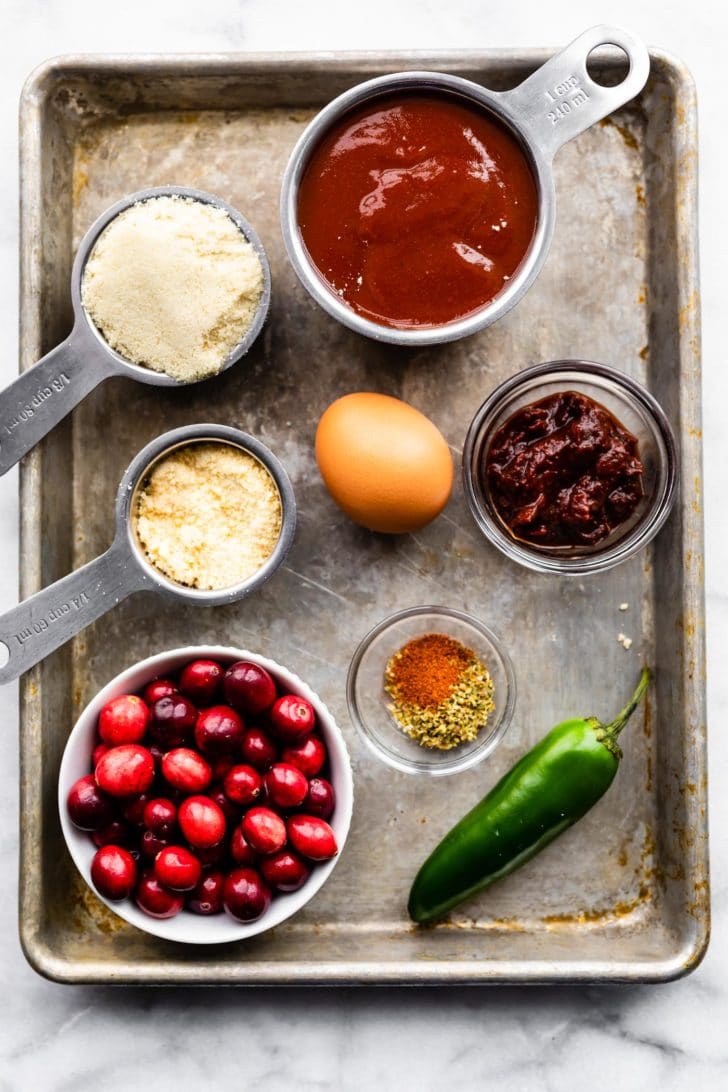 Ingredients for gluten free meatballs with cranberry sauce including Parmesan, almond flour, cranberries, bbq sauce, an egg, peppers in adobo sauce, a jalapeno, and spices