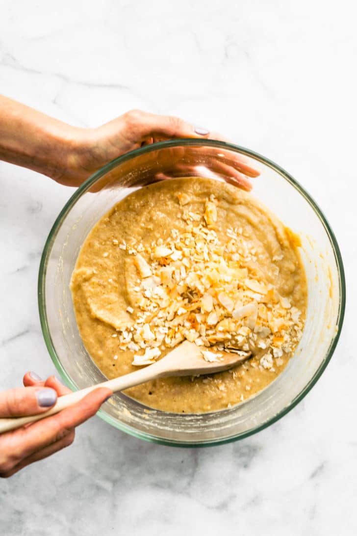 two hands holding a glass bowl using a wooden spoon to mix the batter for almond flour banana muffins