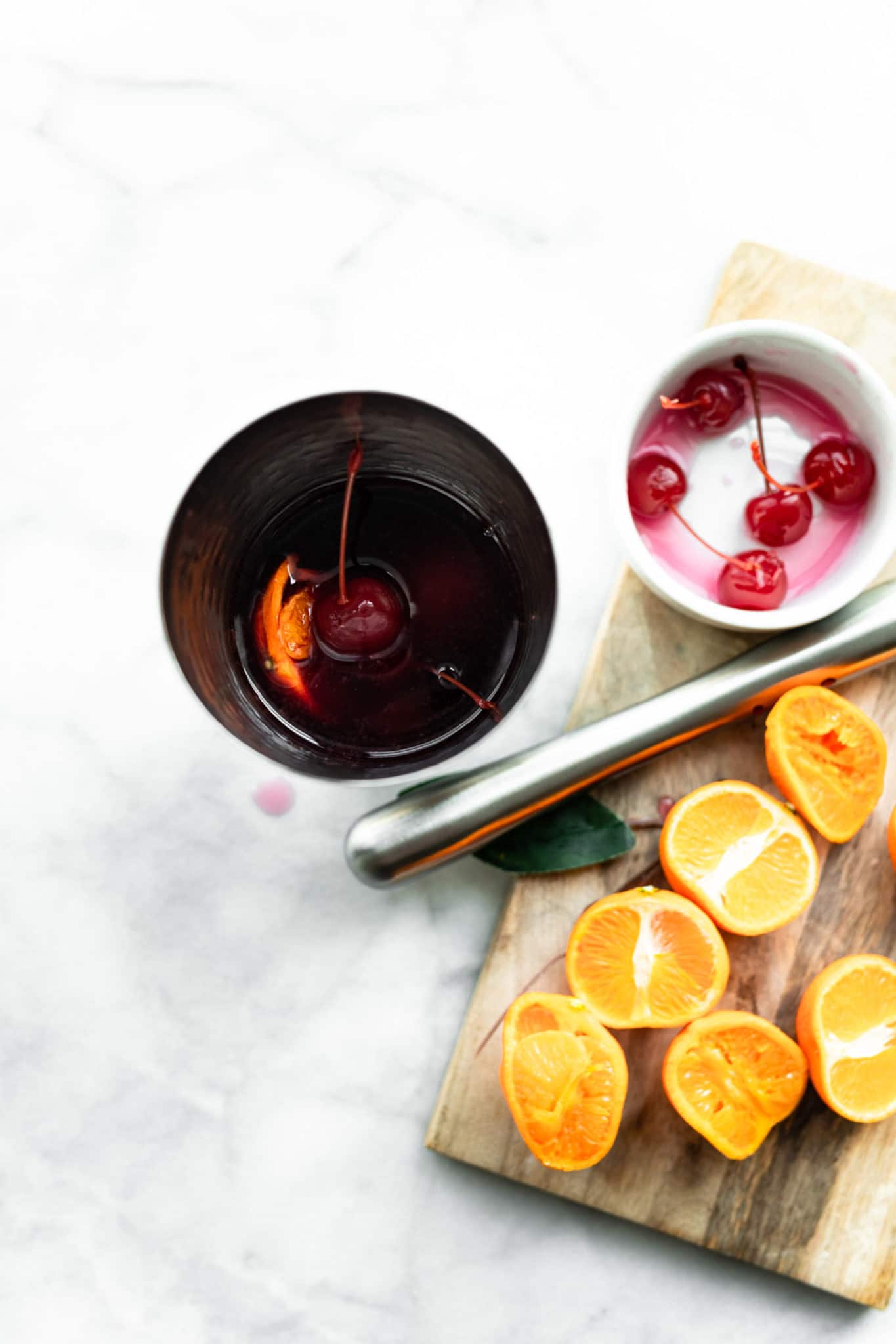 ingredients for a yuletide moon red wine cocktail in a cocktail shaker with a cutting board topped with sliced oranges and a white bowl containing cherries