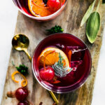 two yuletide moon red wine cocktails on a cutting board topped with an orange slice, ice, and a cherry with fruit and a muddler on the side