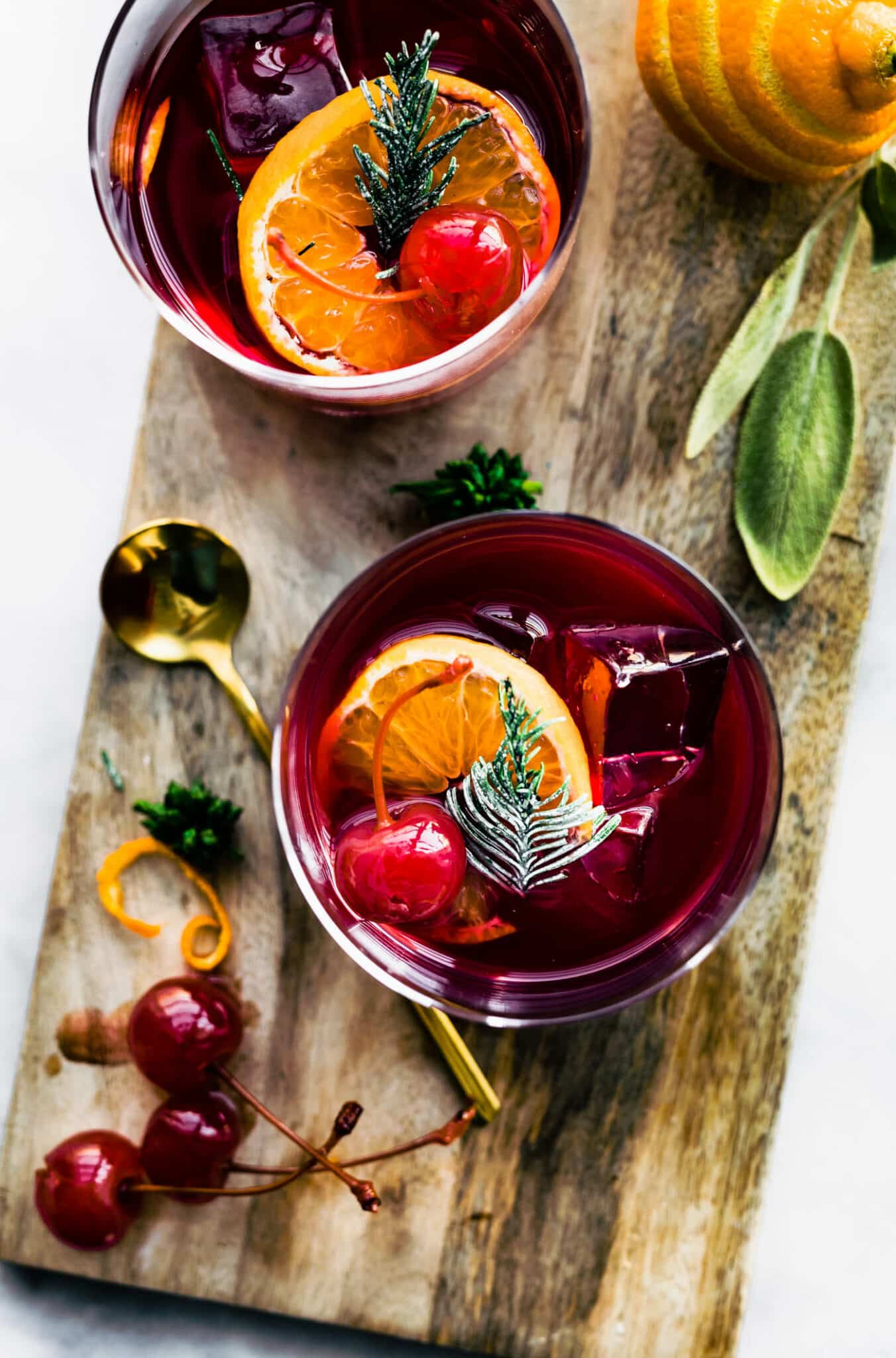 two yuletide moon red wine cocktails on a cutting board topped with an orange slice, ice, and a cherry with fruit and a muddler on the side