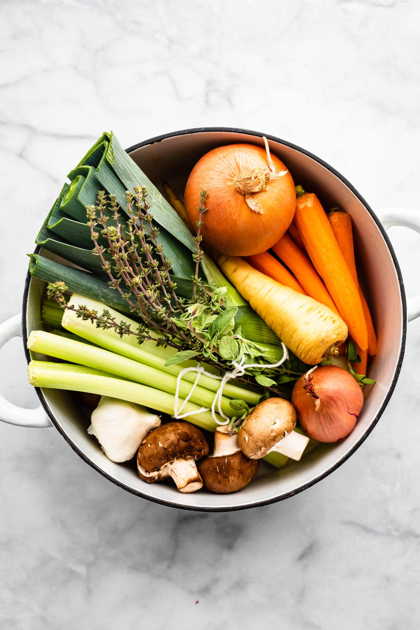 a pan full of vegetables including an onion, shallot, carrots, a parsnip, celery, leaks, mushrooms, and fresh herbs showing how to make vegetable broth
