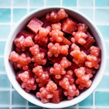 a white bowl on a counter with a blue tiled background filled with red colored homemade gummies with probiotics in the shape of cubes and bears