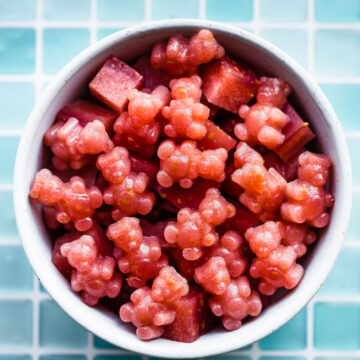 a white bowl on a counter with a blue tiled background filled with red colored homemade gummies with probiotics in the shape of cubes and bears