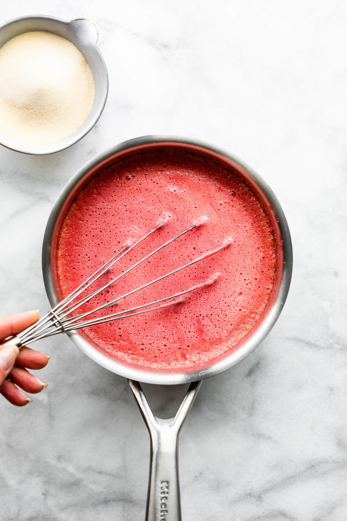 a hand holding a whisk, whisking gelatin powder into a saucepan with blended honey, raspberries, and kombucha for homemade gummies with probiotics