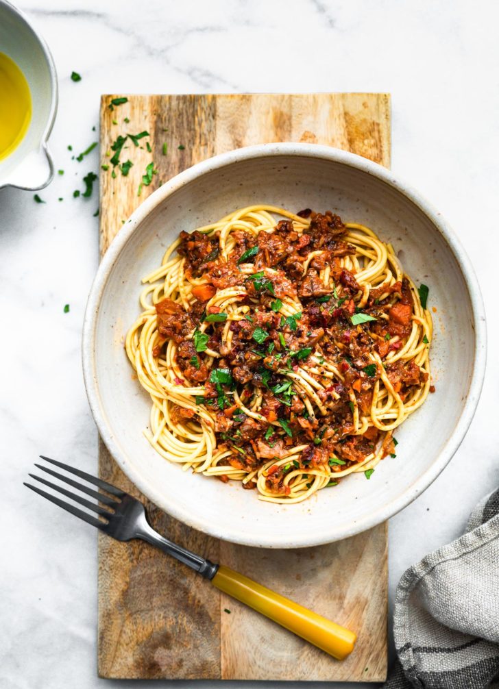 an overhead image of a white bowl full of long gluten-free pasta noodles topped with vegan muhsrrom bolognese, grated cheese, and fresh herbs with a fork on the side