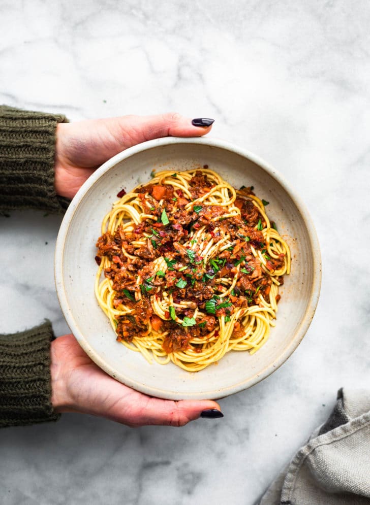 overhead image of two hands holding a white bowl full of gluten-free pasta noodles topped with vegan mushroom bolognese, grated cheese, and fresh herbs