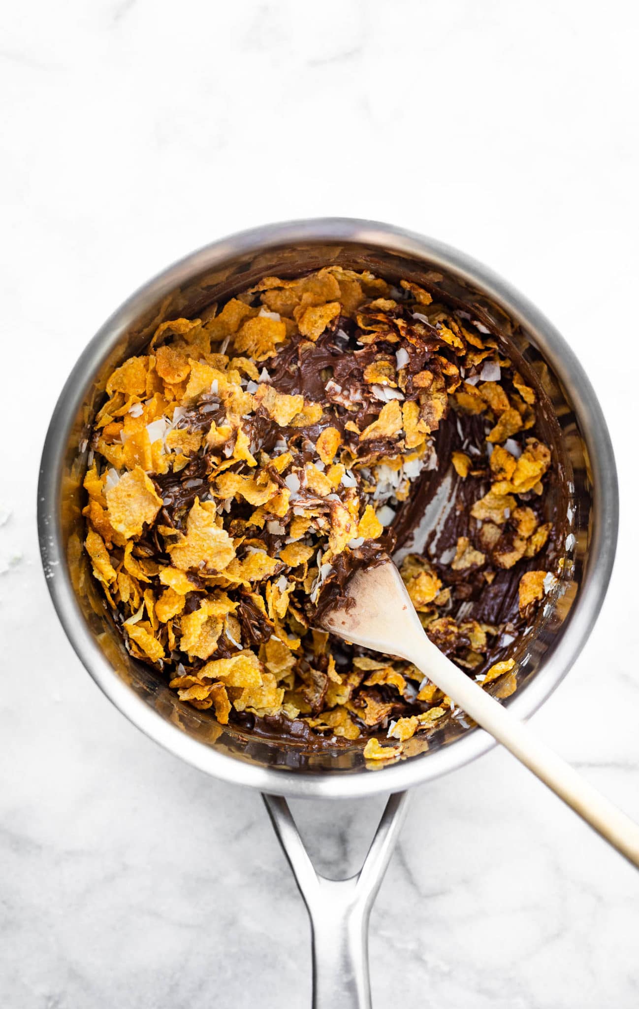 a wooden spoon in a saucepan stirring cornflake cereal into a chocolate peanut butter mixture to create chocolate peanut butter cornflake cookies