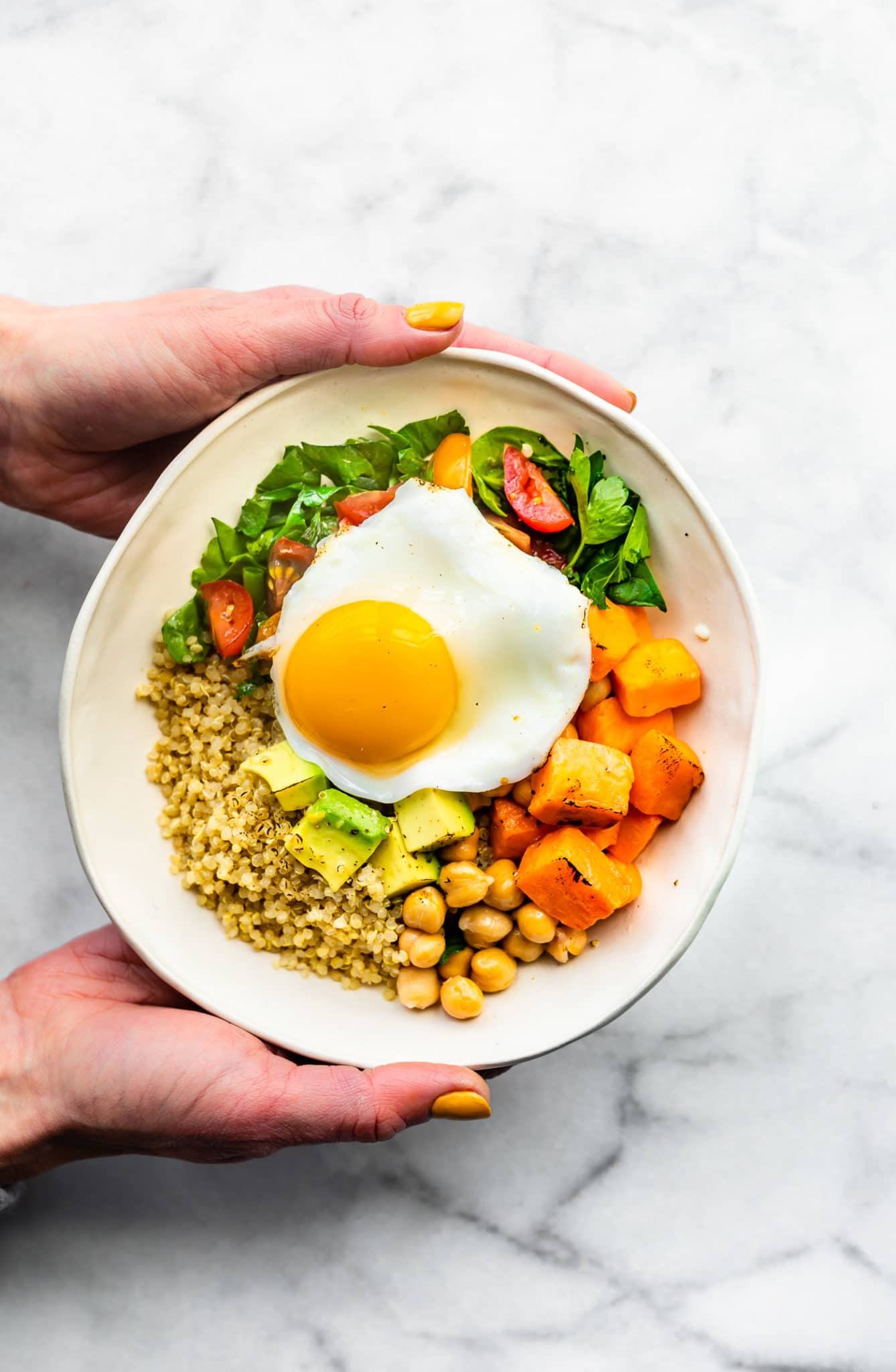 two hands holding a white savory quinoa breakfast bowl with quinoa, chickpeas, roasted sweet potatoes, spinach, tomatoes, avocado, and a fried egg