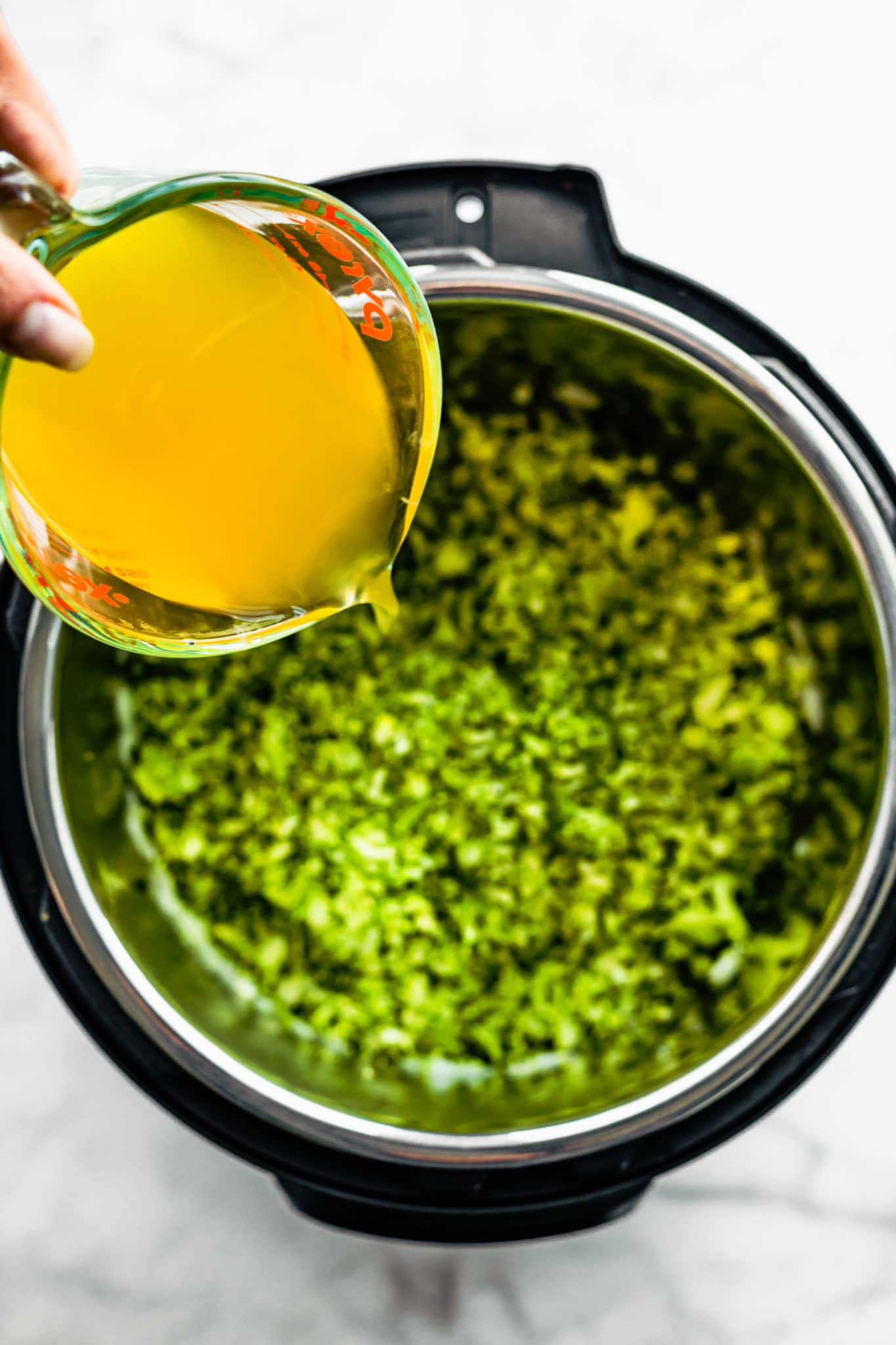 overhead image of a hand pouring veggie broth into an instant pot with diced broccoli inside