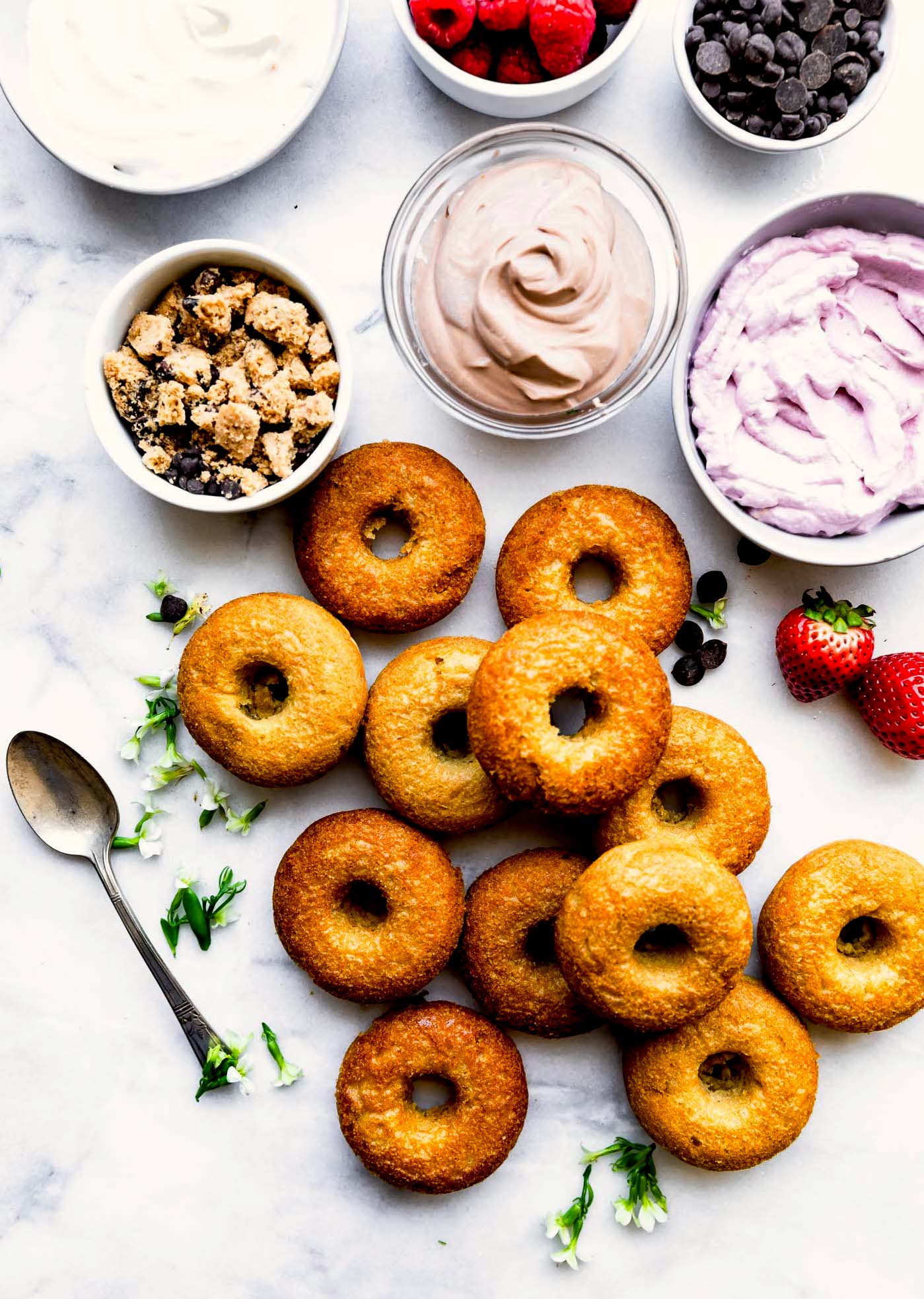 unglazed baked vegan donuts with bowls of frosting including a chocolate flavor, a berry flavor, and a vanilla flavor with bowls of cookie pieces, chocolate chips, and raspberries