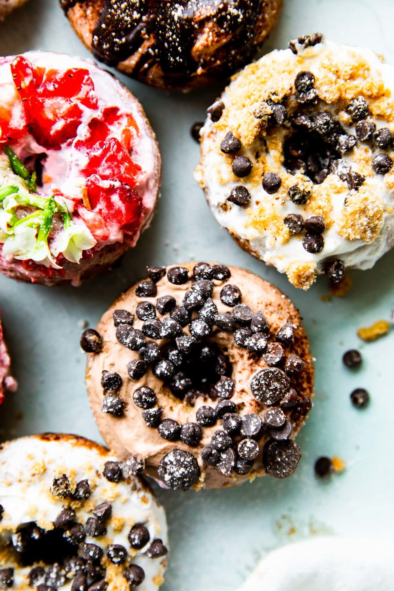 a close up image of vanilla, chocolate, and vanilla vegan baked donuts topped with frosting, chocolate chips, and fruit