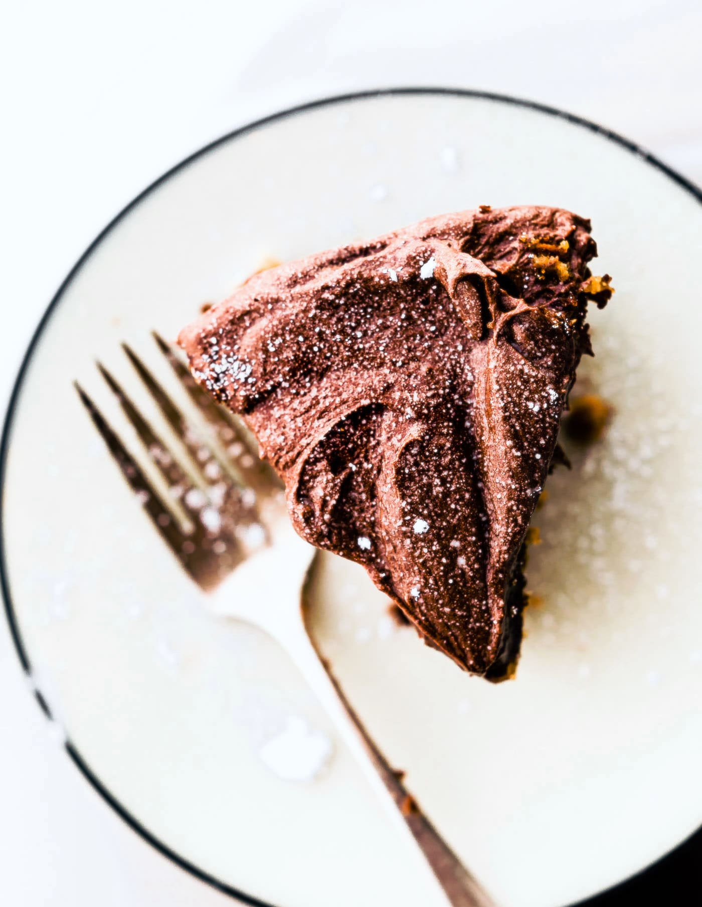 a piece of vegan vanilla cake with chocolate frosting on a white plate with a fork on the side