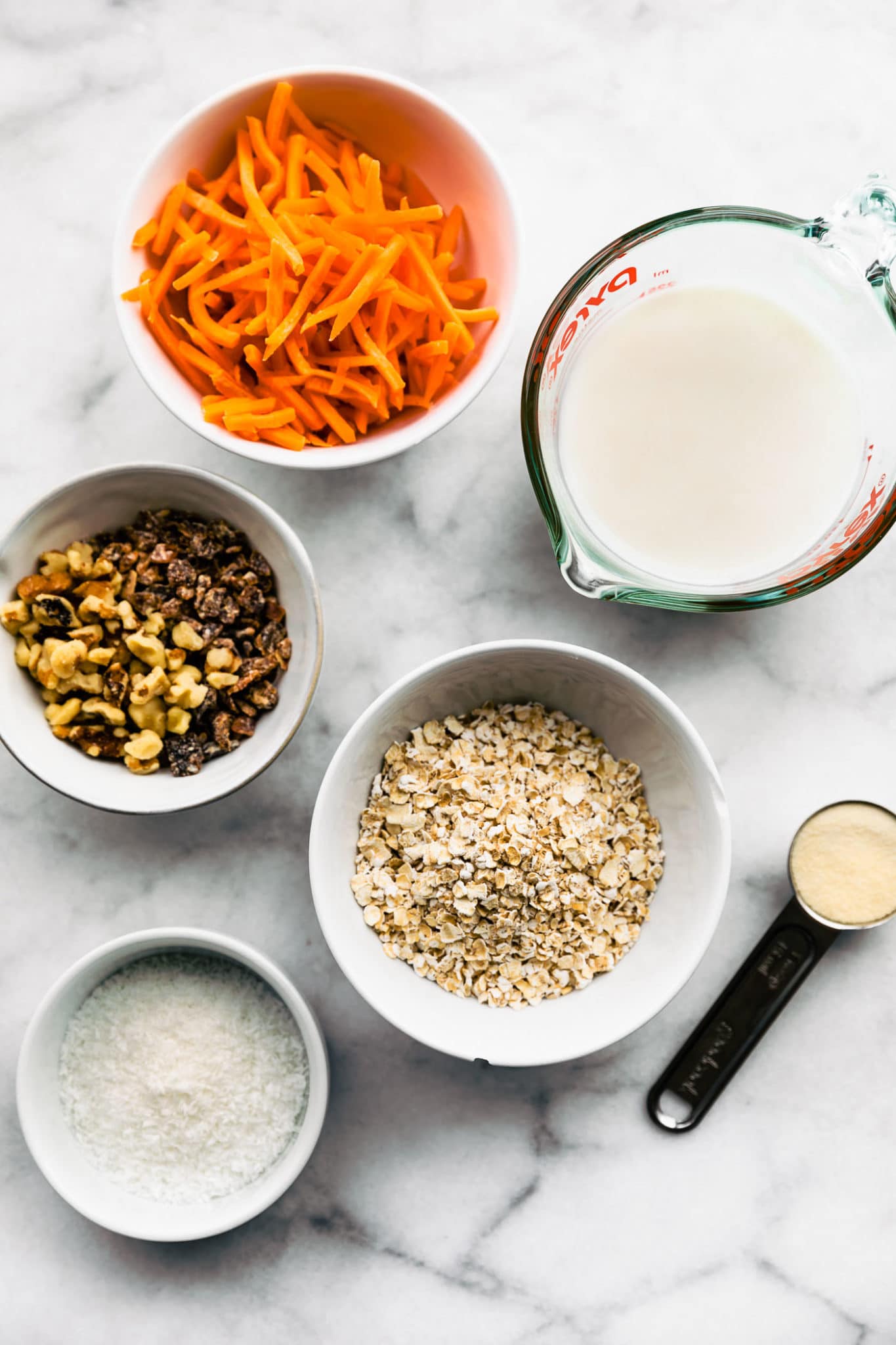 bowls of ingredients containing items for healthy carrot cake muffins including shredded carrots, chopped walnuts, raisins, gluten free oats, dairy-free milk, and sugar