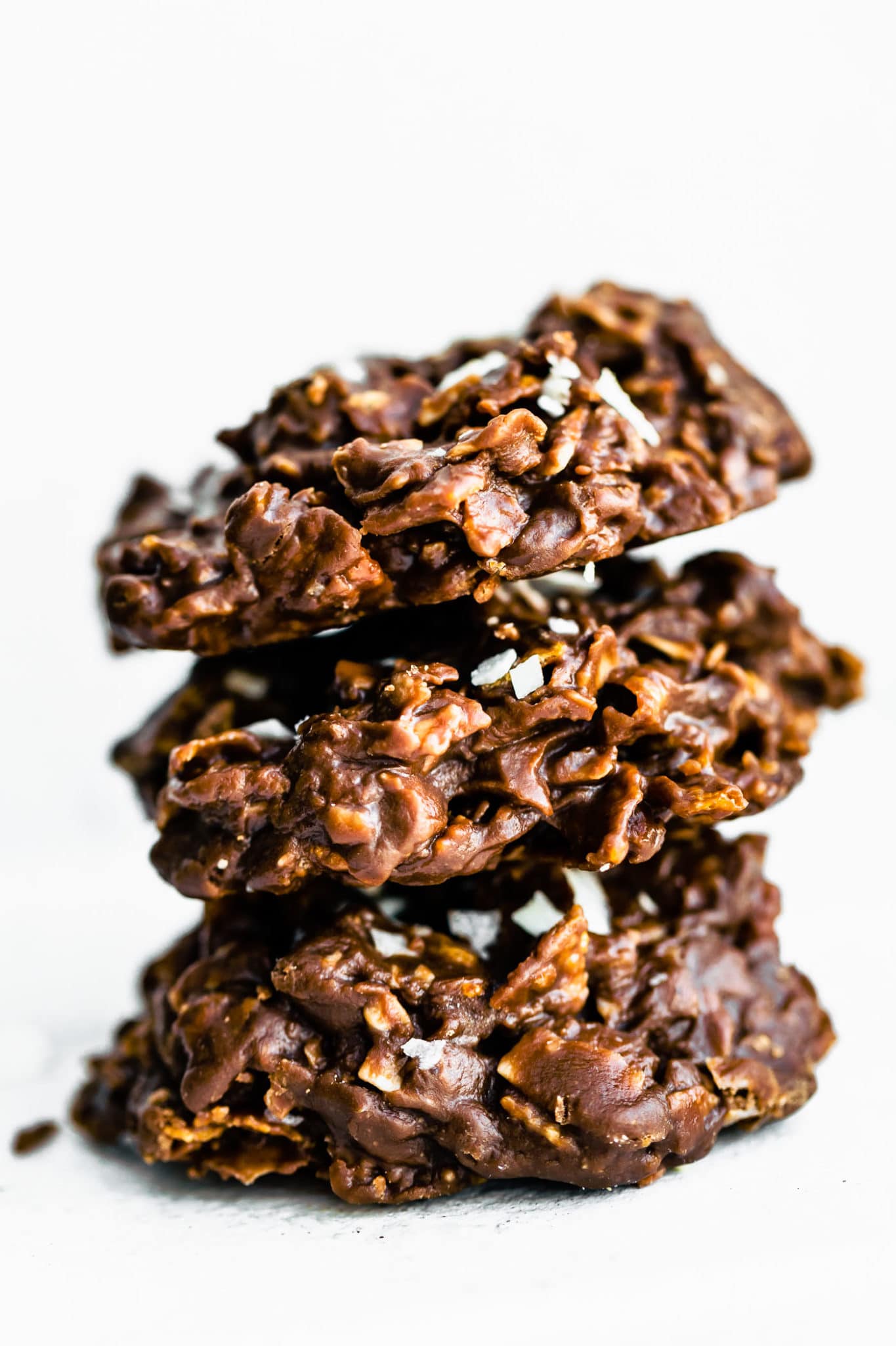 3 no-bake chocolate peanut butter cornflake cookies topped with coconut flakes and sea salt stacked on top of each other