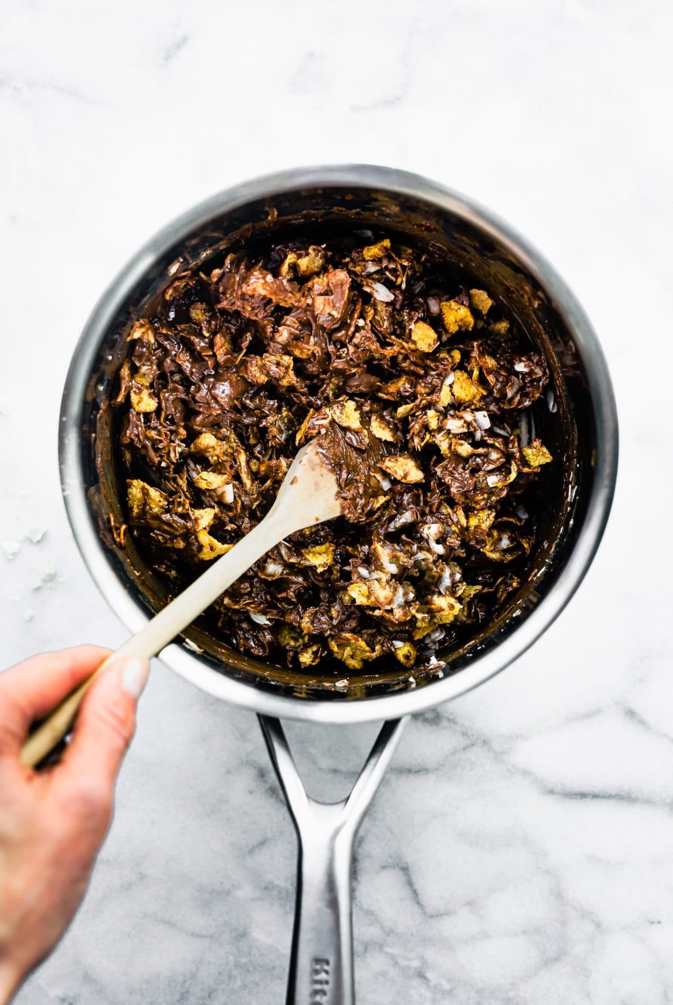 a woman's hand holding a wooden spoon in a saucepan folding cornflake cereal into a chocolate peanut butter mixture to make chocolate peanut butter cornflake cookies
