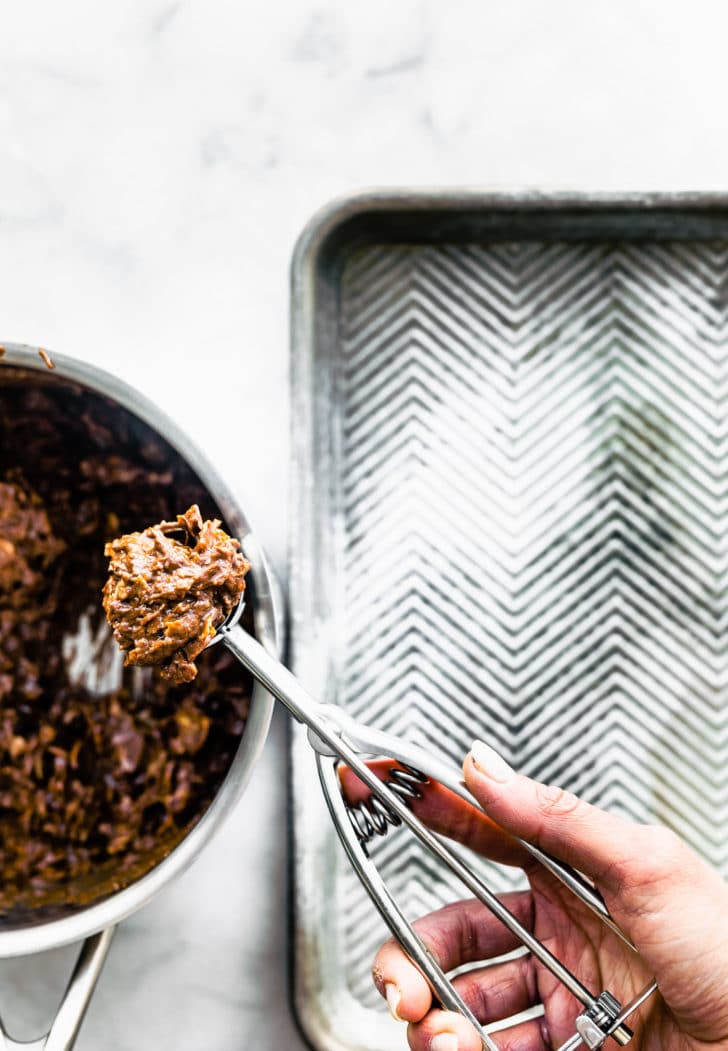 a bowl of chocolate peanut butter cornflake cookie batter next to a baking dish with a woman's hand holding a cookie scoop full of batter over the bowl