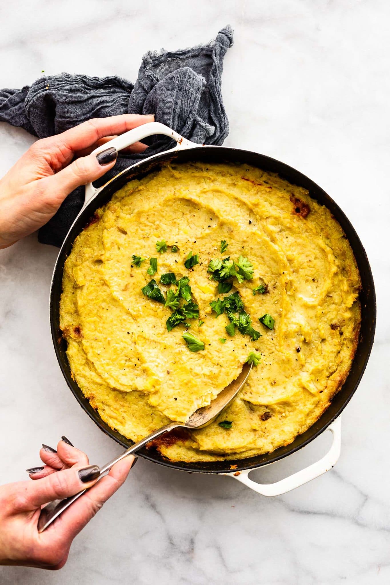 a woman's hands dipping a spoon into a pan full of potato and cauliflower shepherd's pie