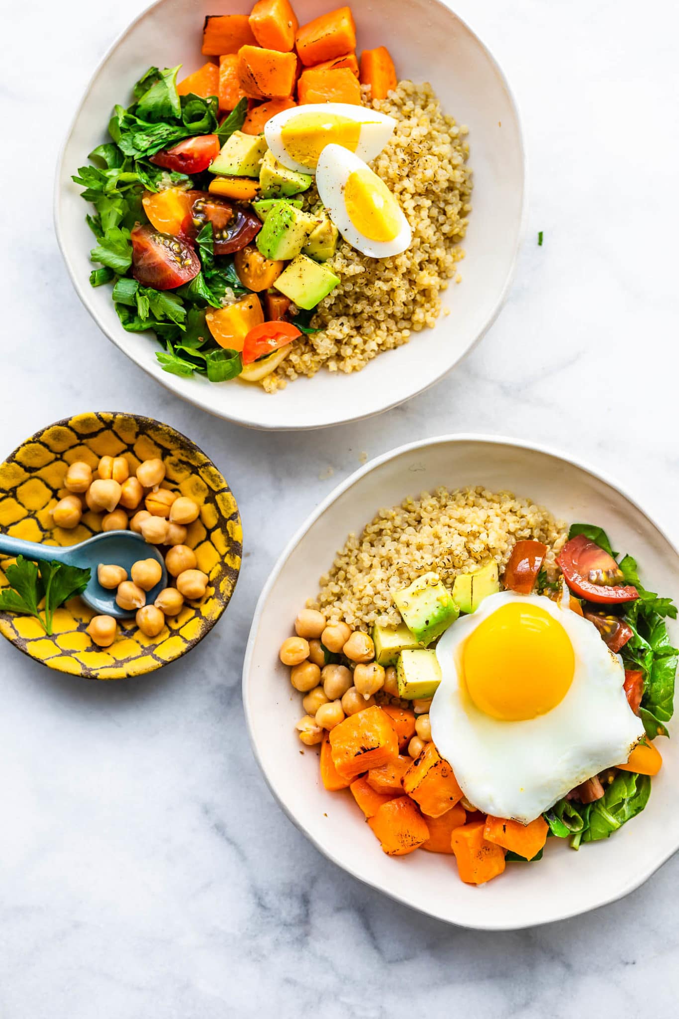 two savory quinoa breakfast bowls one containing quinoa, sweet potatoes, tomatoes, spinach, avocado, and a boiled egg the other containing quinoa, chickpeas, sweet potato, tomatoes, spinach, and a fried egg, and a small bowl with chickpeas on the side