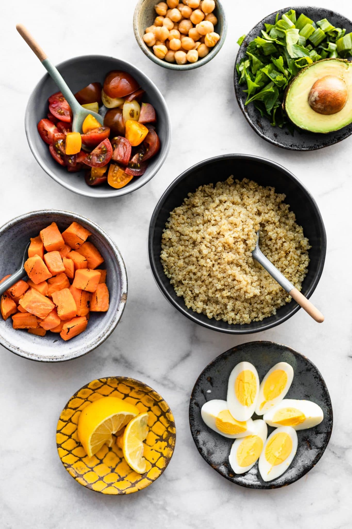 bowls containing ingredients for savory quinoa breakfast bowls including lemon wedges, boiled eggs, quinoa with a serving spoon, roasted sweet potatoes with a serving spoon, sliced tomatoes with a serving spoon, chickpeas, spinach, and half an avocado