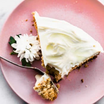 a slice of vegan vanilla cake with vanilla frosting on a light pink plate with a fork breaking off a piece and a white edible flower on the side