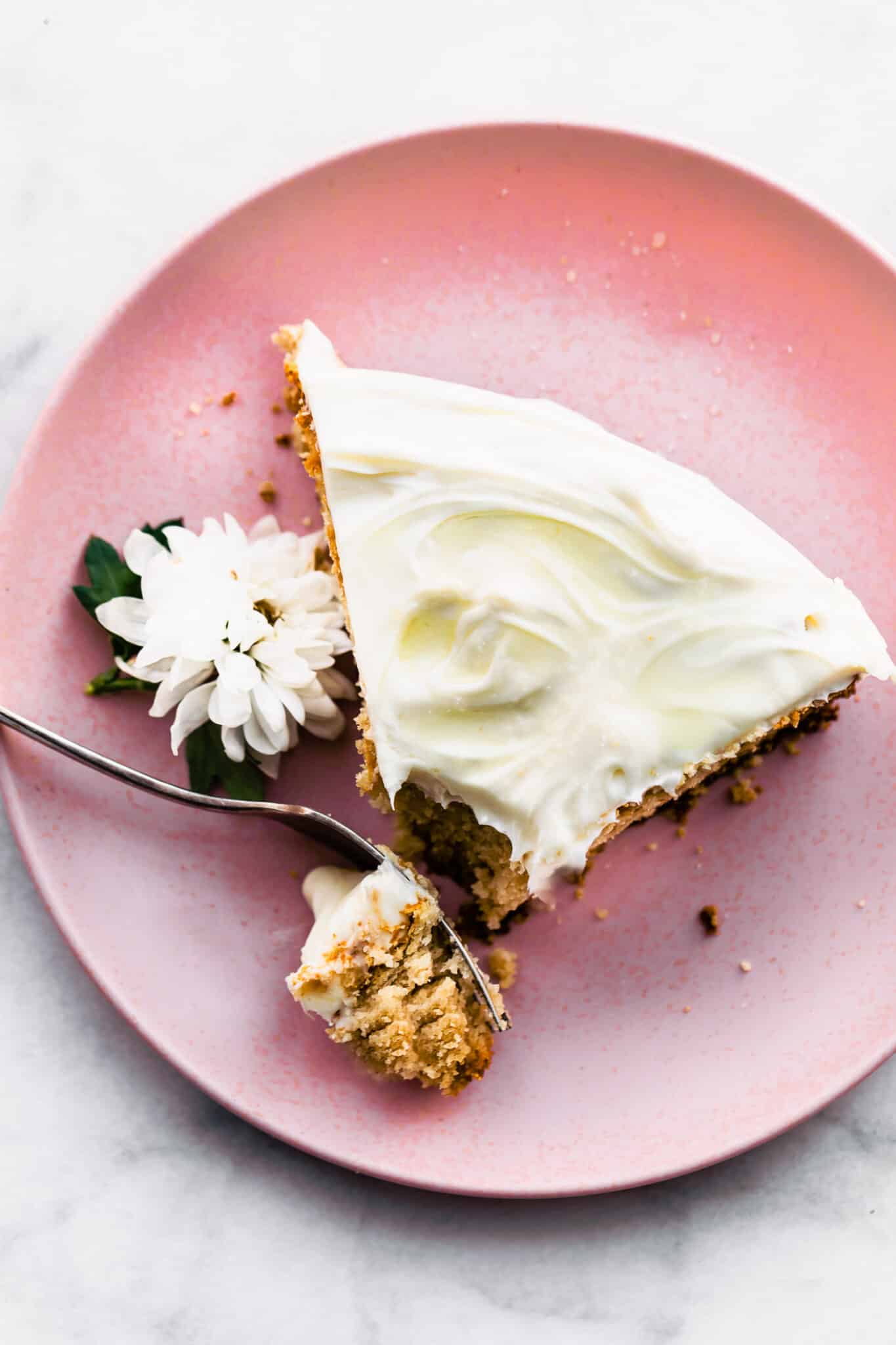 a slice of vegan vanilla cake with vanilla frosting on a light pink plate with a fork breaking off a piece and a white edible flower on the side