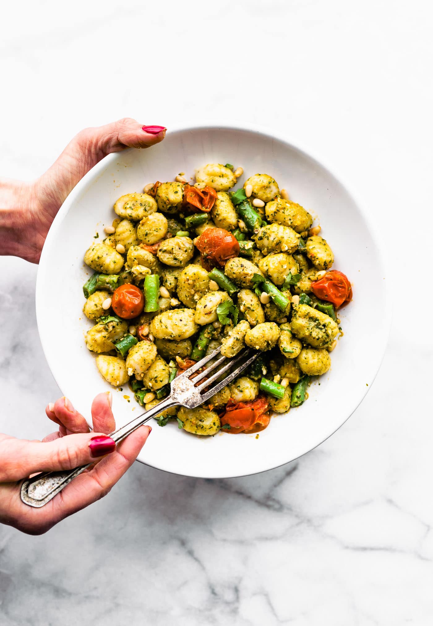 two hands holding a white bowl full of gluten free gnocchi pesto pasta salad with blistered cherry tomatoes and green beans and a fork