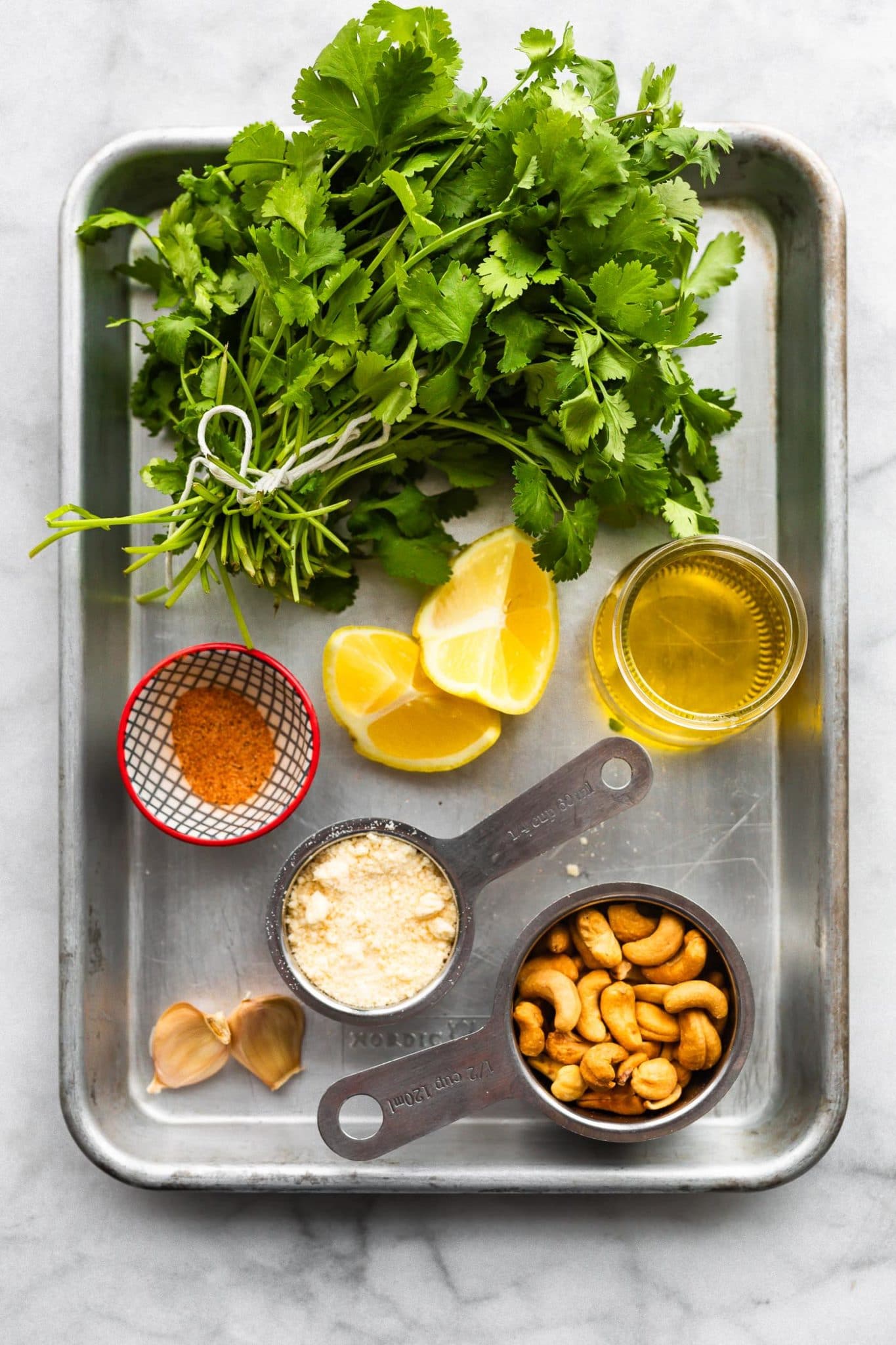 a baking pan willed with ingredients for cilantro pesto including cilantro, lemon wedges, olive oil, spices, grated Parmesan, cashews, and garlic