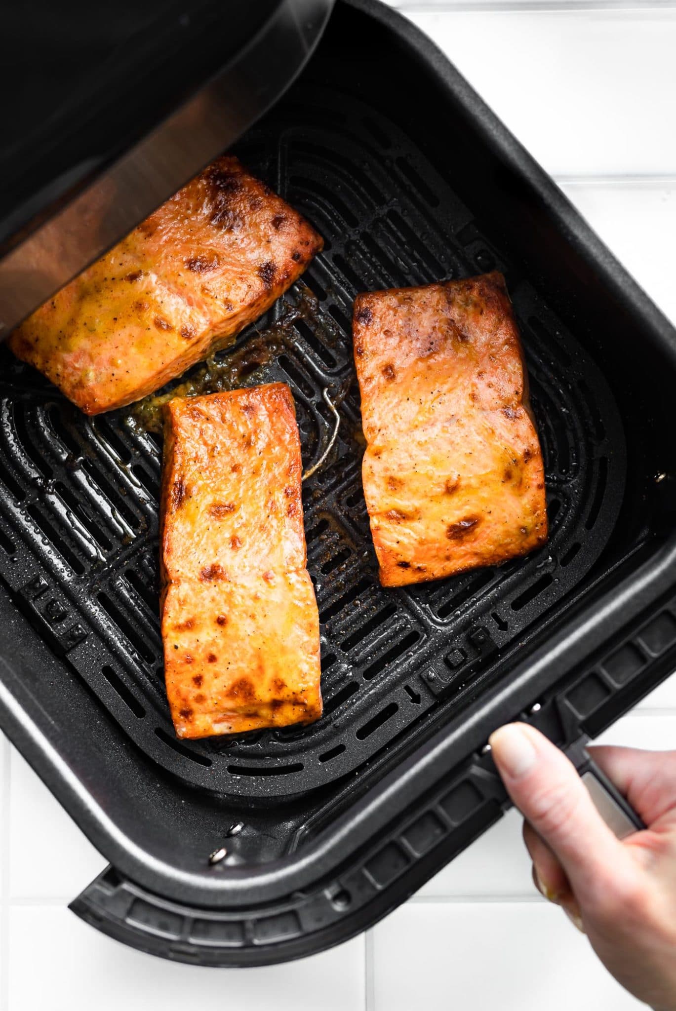 a woman's hand holding the handle of an air fryer basket that is filled with 3 pieces of cooked air fryer salmon topped with creamy mustard sauce