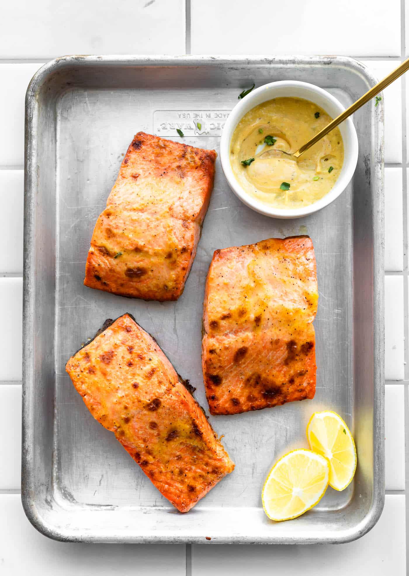 three pieces of air fried salmon in a baking dish topped with mustard sauce with lemon wedges and a white bowl of mustard sauce with a yellow spoon dipped in on the side