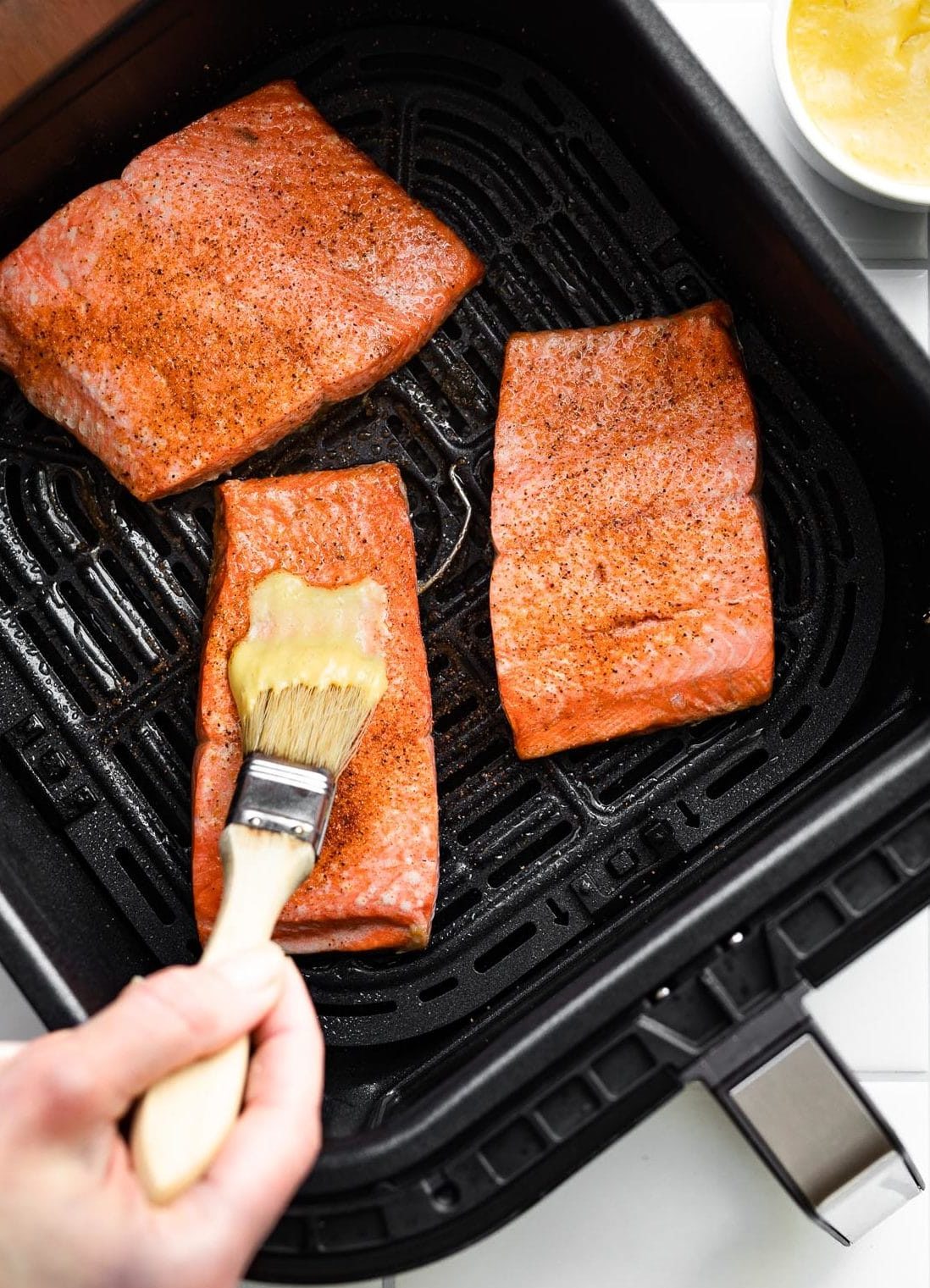 3 pieces of seasoned salmon in the basket of an air fryer with a small white bowl full of creamy mustard sauce on the side and a woman's hand holding a brush spreading the mustard sauce on top of one of the salmon filets