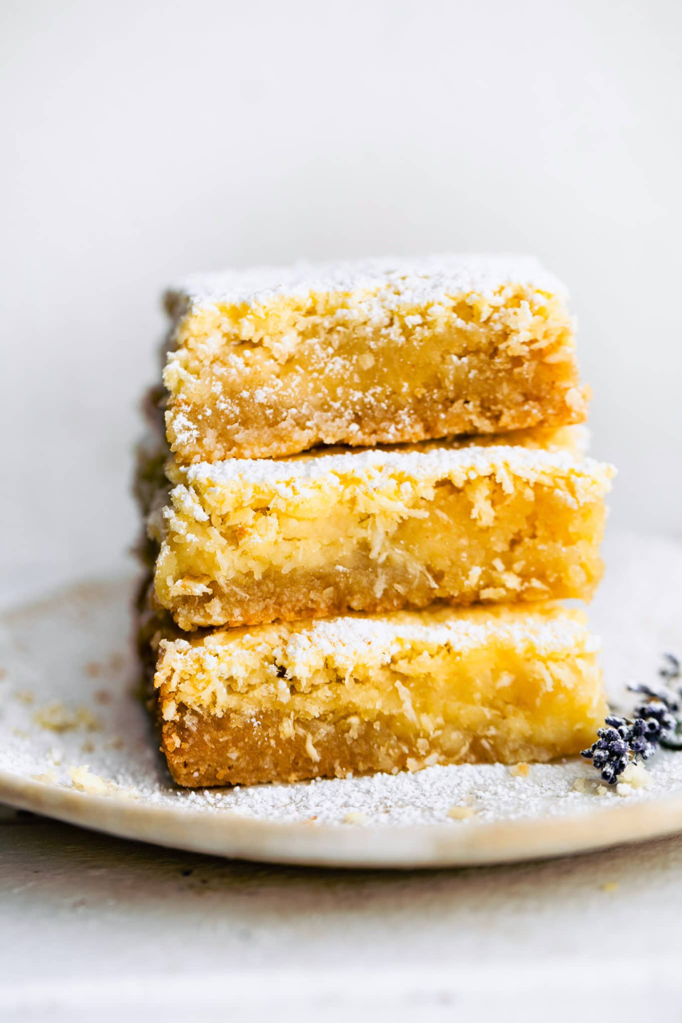 a stack of three gluten free lemon bars stacked on top of each other on a white plate with a sprig of lavendar on the side