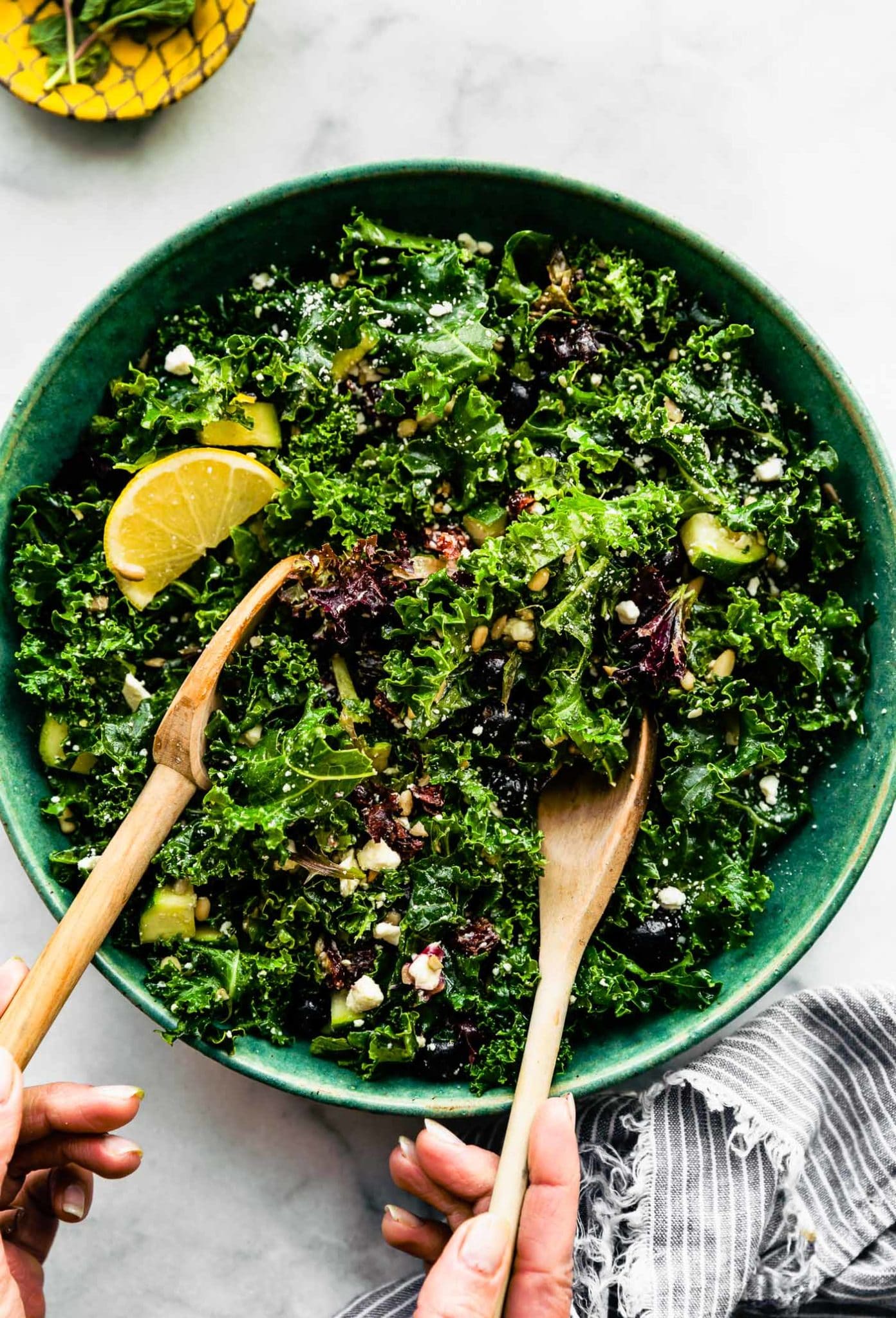 two hands using wooden spoons to toss massaged kale salad in a bowl topped with a lemon wedge