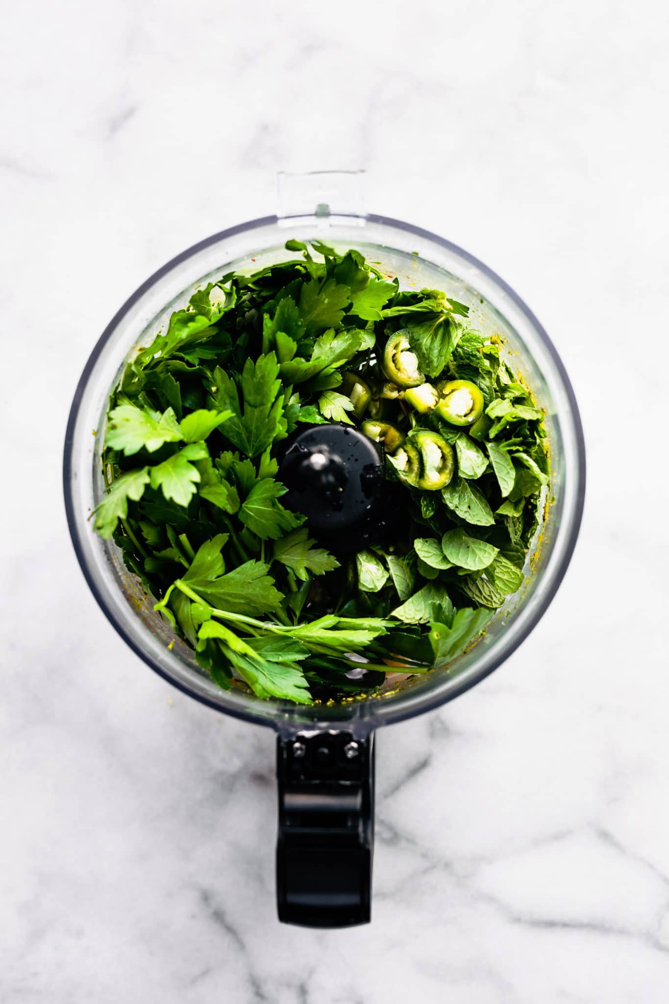 overhead image of a food processor on a white marble countertop filled with fresh cilantro, mint, peppers, and spices to create a mint chimichurri sauce recipe