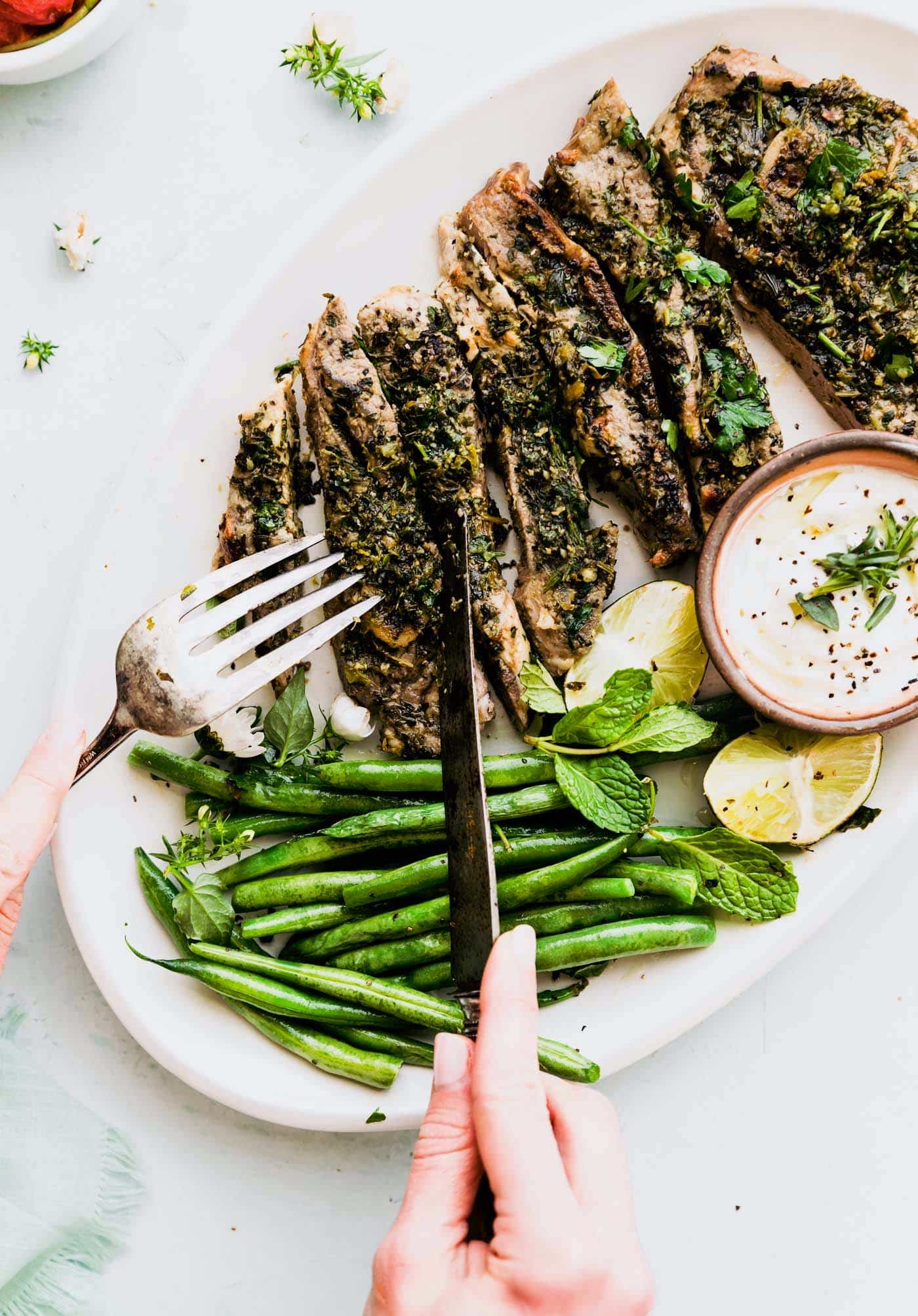 a white serving platter with broiled lamb chops with mint chimichurri sauce and green beans, lime wedges, and a white dipping sauce on the side with two hands holding a fork and a knife ready to cut into a lamb chop