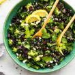 a large blue bowl filled with massaged kale salad topped with lemon wedges and two wooden serving spoons