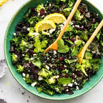 a large blue bowl filled with massaged kale salad topped with lemon wedges and two wooden serving spoons