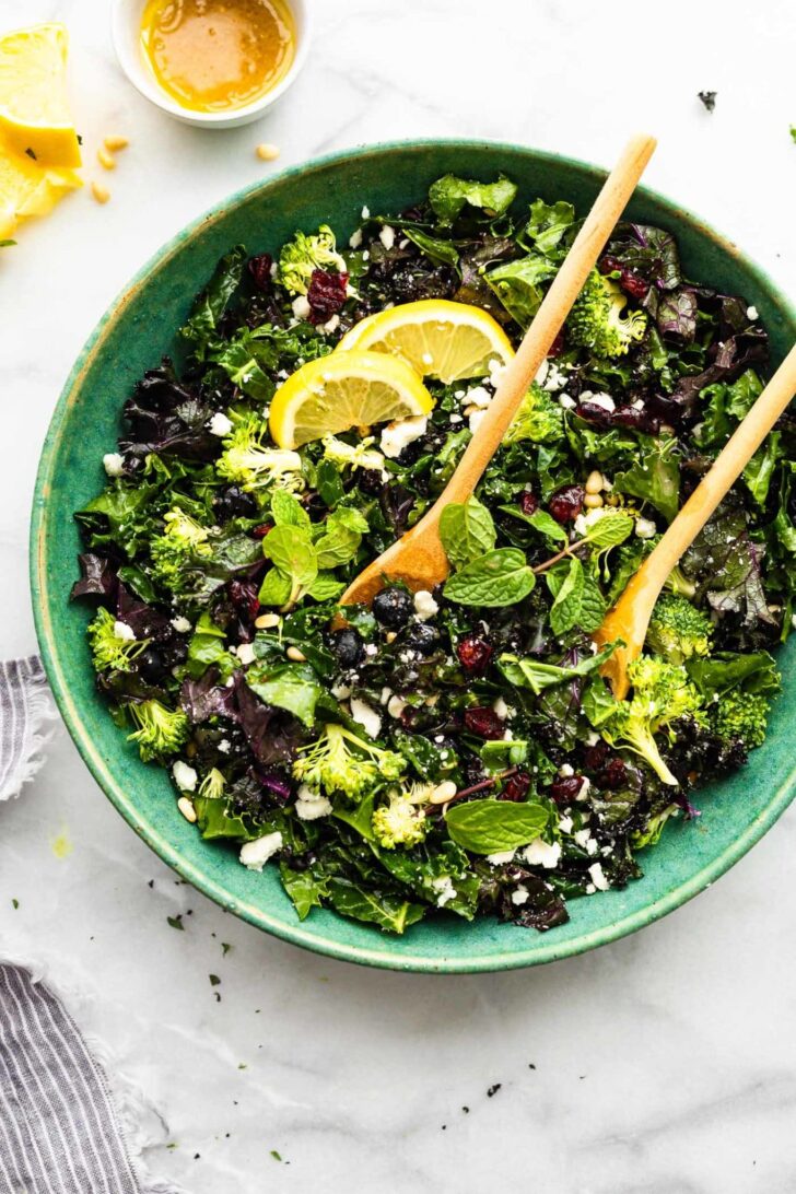 A large blue bowl filled with massaged kale salad topped with lemon wedges and two wooden serving spoons.
