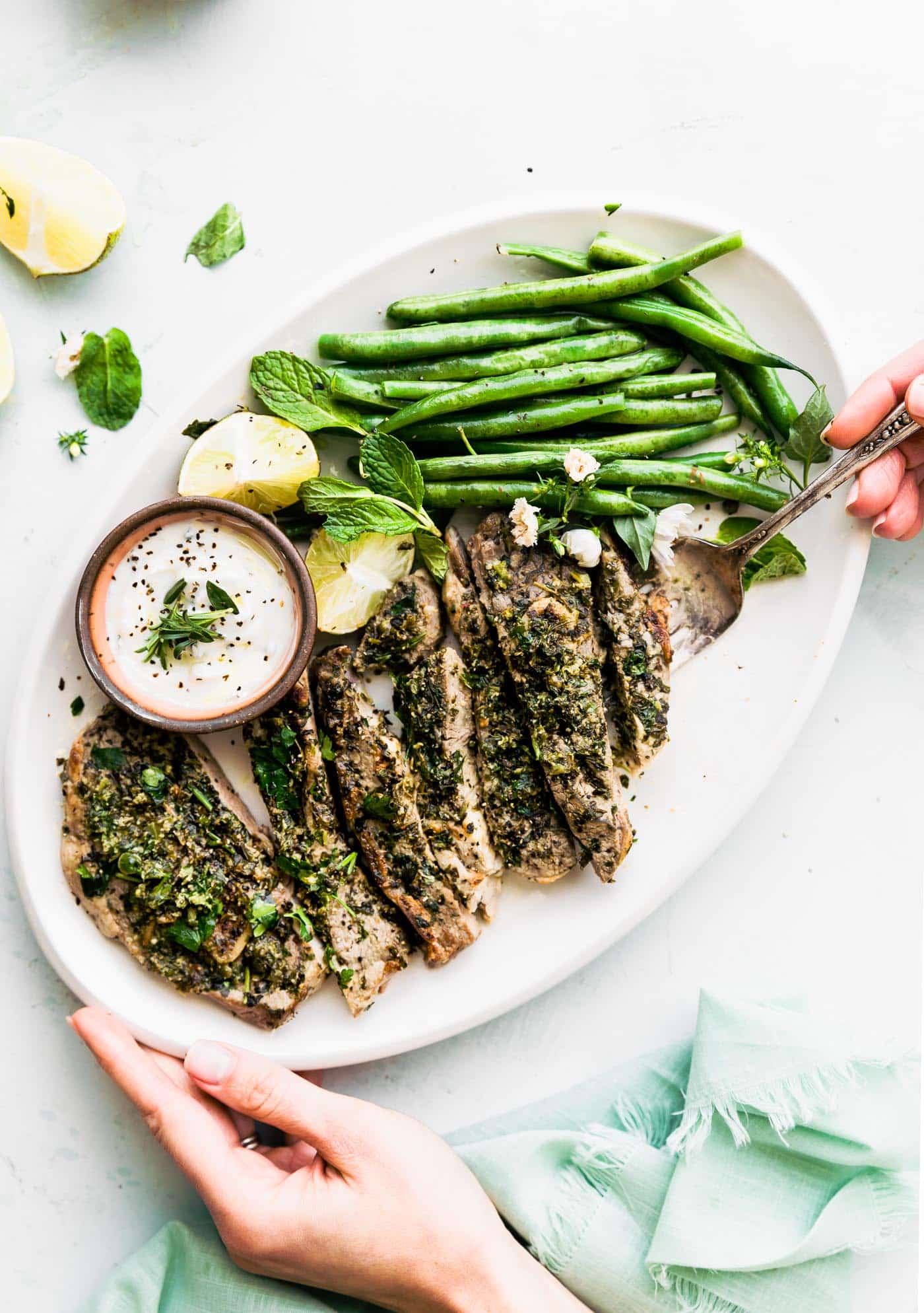 a white serving platter filled with broiled lamb chops with mint chimichurri sauce, green beans, a halved lime, and a white yogurt dipping sauce with a woman's hand holding the platter and another hand holidng a fork underneath a lamb chop