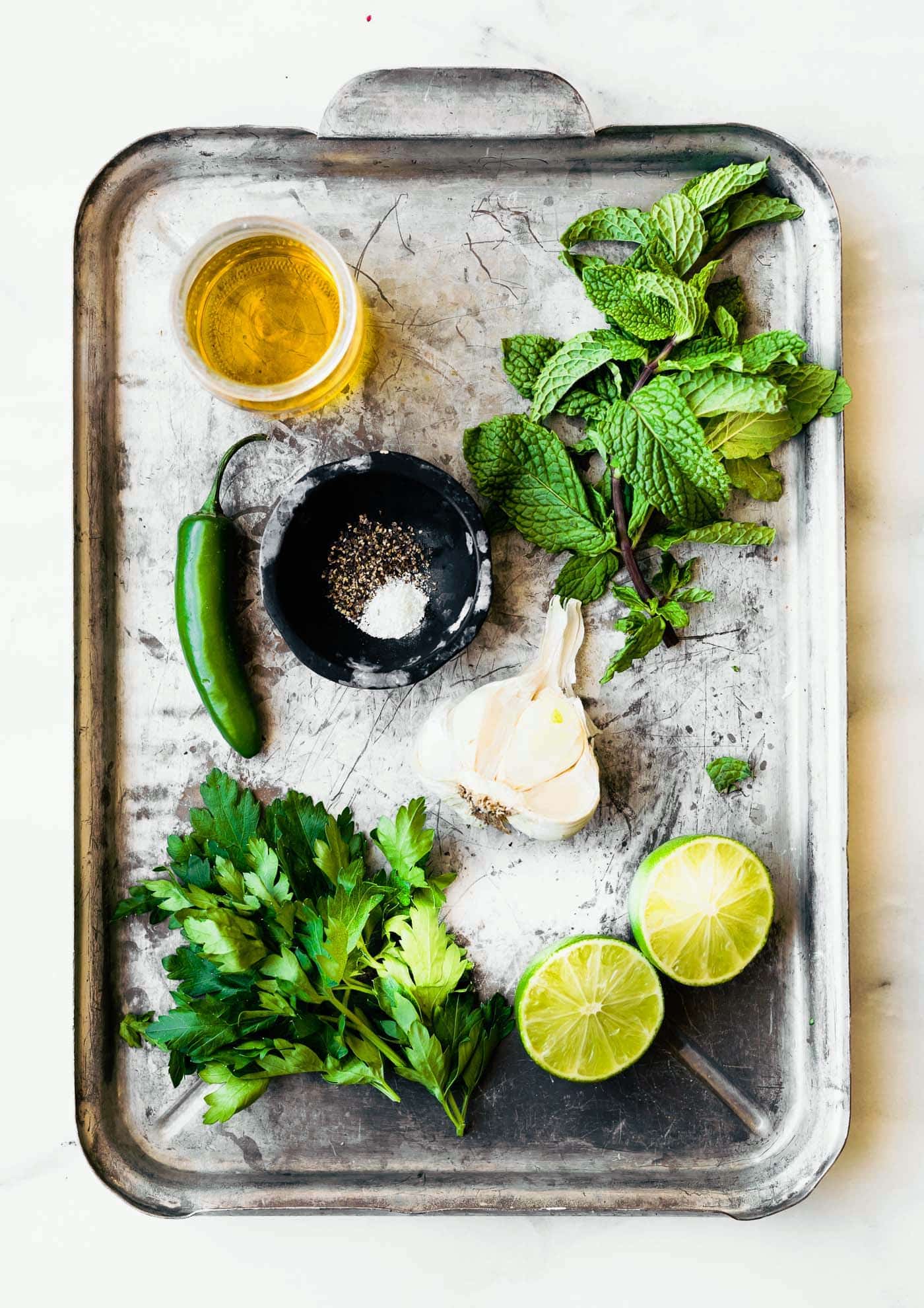 a serving tray with a cup of olive oil, fresh parsley, fresh mint, a jalapeno pepper, a black bowl with salt and pepper, garlic cloves, and a halved lime