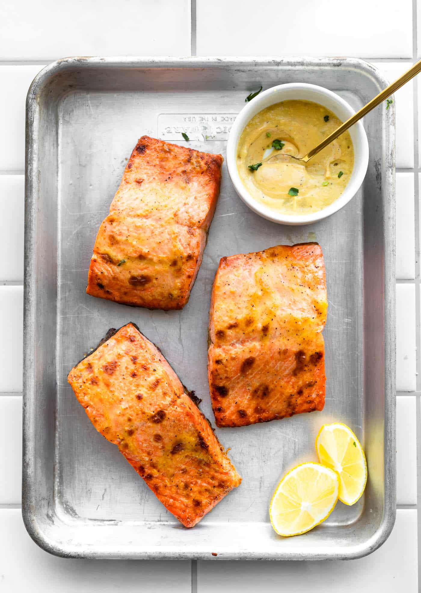 3 pieces of air fryer salmon in a baking dish with lemon wedges and a white bowl filled with creamy mustard sauce on the side