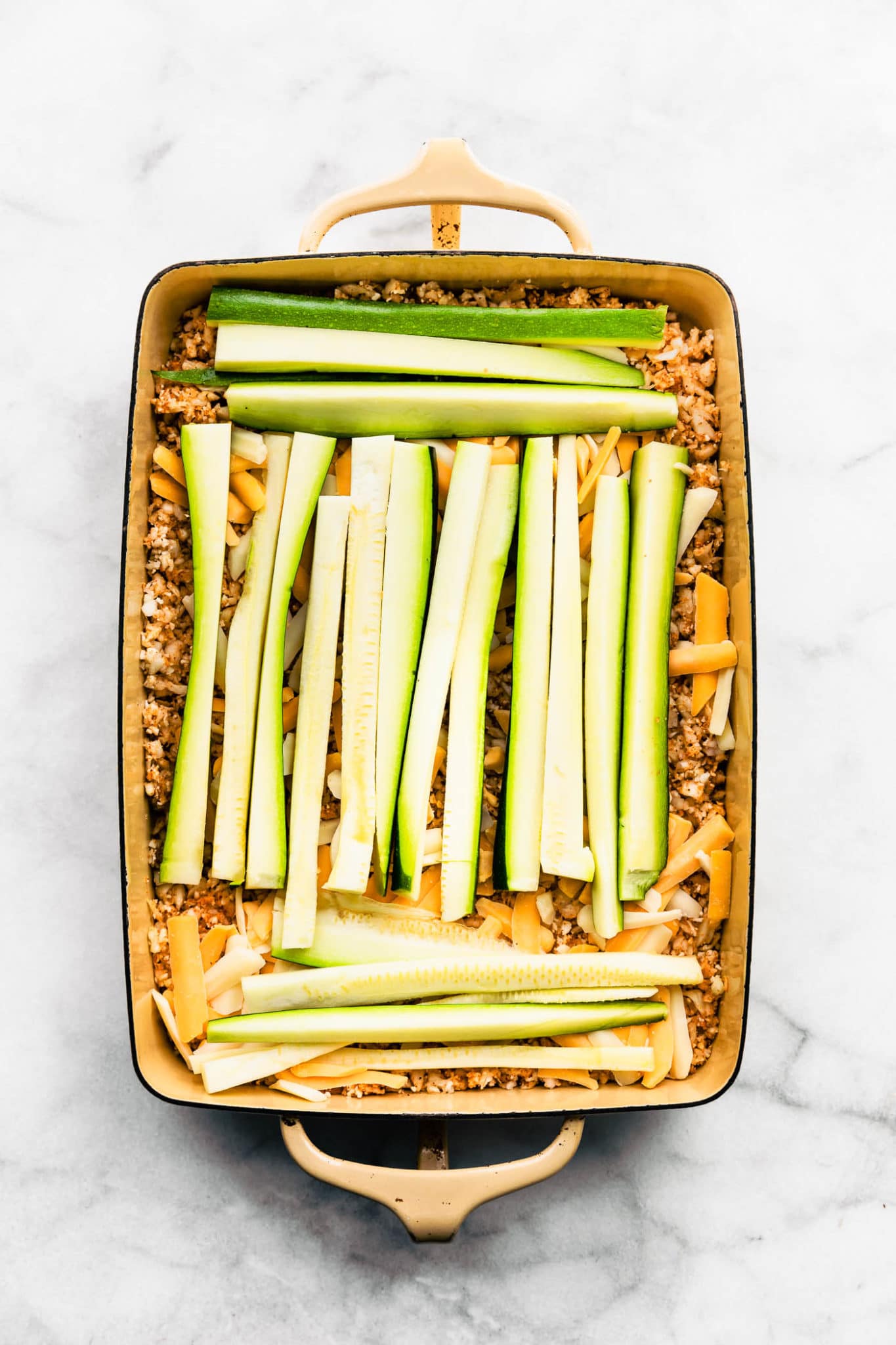 overhead image of zucchini strips layered on top of cheese and cauliflower in a casserole dish for tex-mex egg and cauliflower casserole