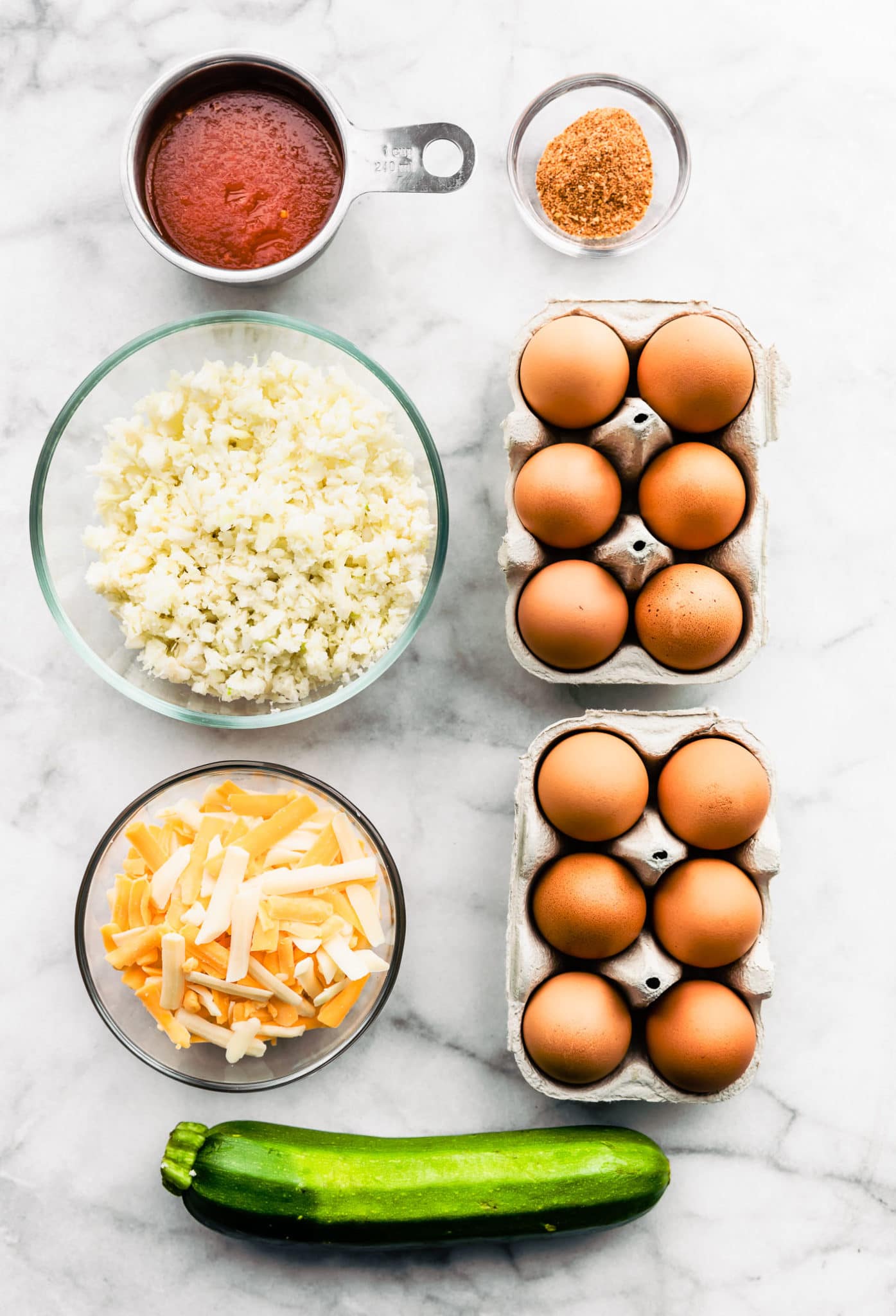 overhead image of two cartons of 6 eggs, a glass bowl with spices, a measuring cup holding enchilada sauce, a glass bowl holding riced cauliflower, a glass bowl holding shredded cheese, and a whole zucchini needed to make tex-mex egg and cauliflower casserole