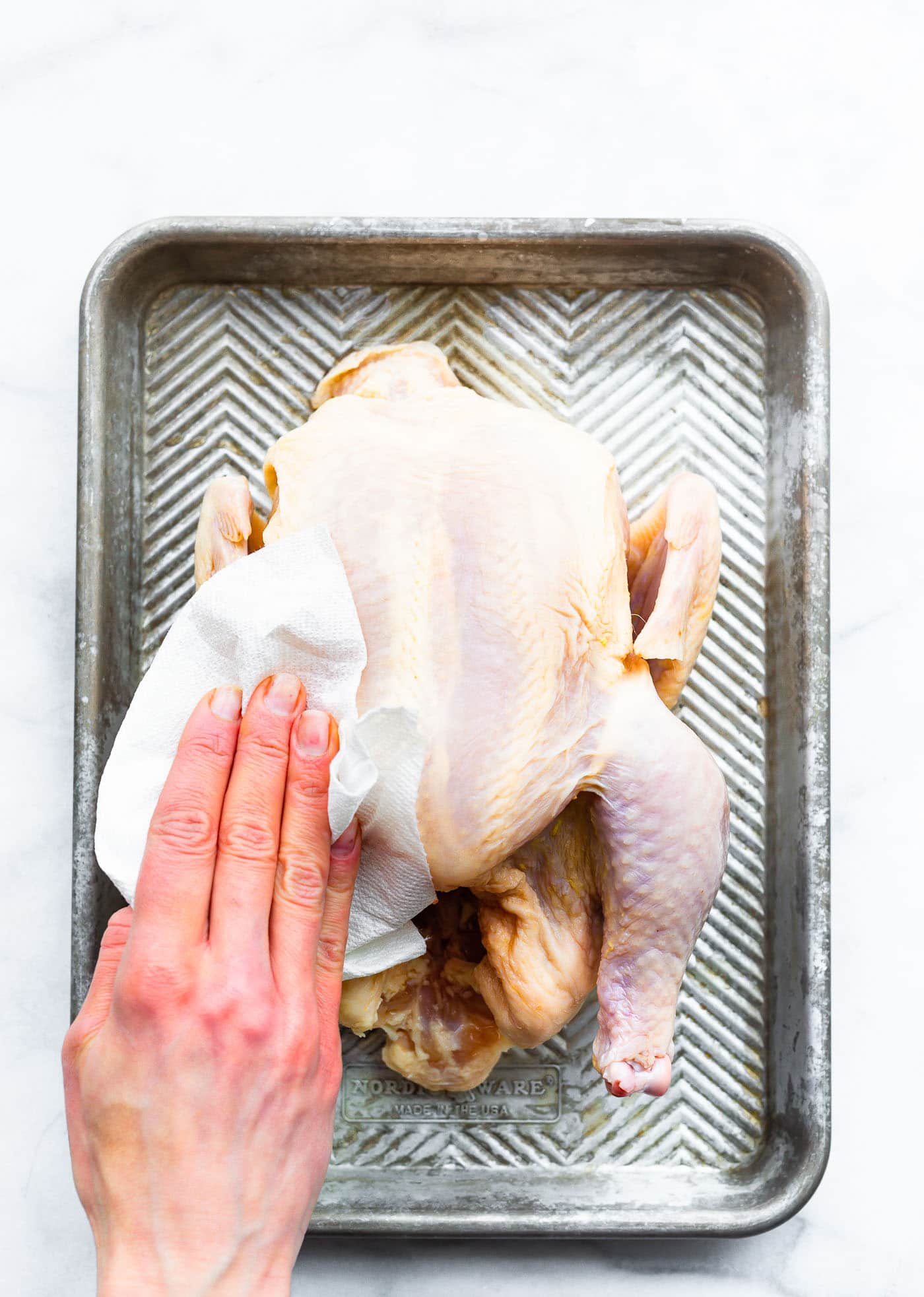 a whole chicken sitting on a baking sheet with a woman's hand using a paper towel to dry any moisture from the skin